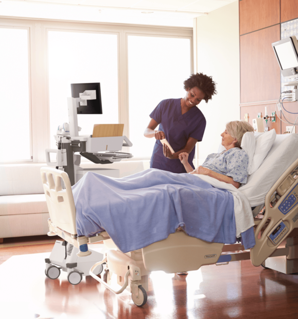 Eine Krankenschwester in violetter Arbeitskleidung unterhält sich mit einer älteren Patientin, die in einem Krankenhausbett liegt, und hält ihre Hand. Der Raum ist hell, da Tageslicht durch große Fenster hereinfällt, und in der Nähe stehen medizinische Geräte.