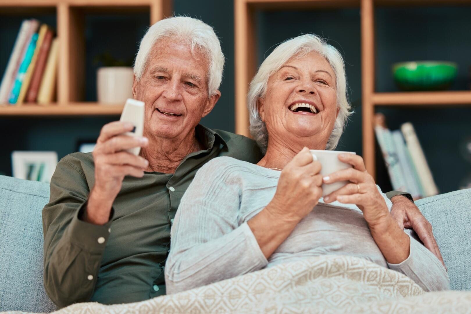 Ein älteres Ehepaar sitzt auf einem Sofa, lächelt und lacht. Der Mann hält eine Fernbedienung in der Hand, die Frau einen weißen Becher. Im Hintergrund sind Bücherregale zu sehen.
