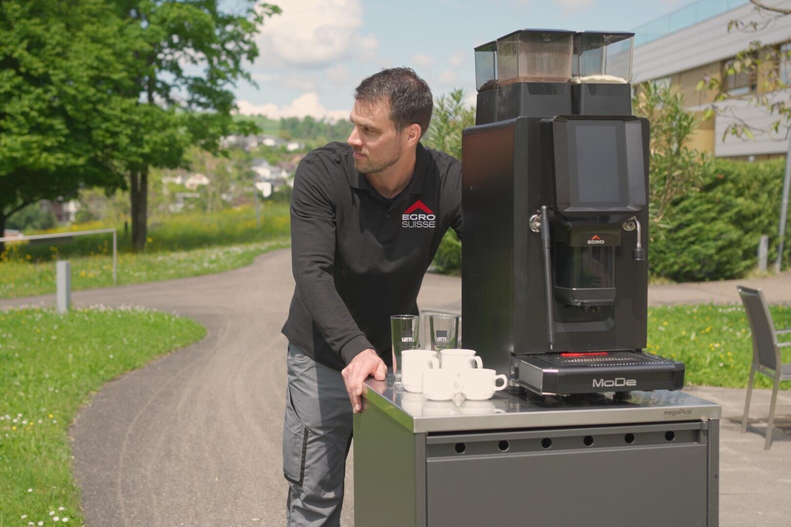 Ein Mann in einem schwarzen Hemd steht draußen neben einer mobilen Kaffeemaschine auf einem Wagen, auf dem Tassen und Gläser stehen. Im Hintergrund sind ein gepflasterter Weg, grünes Gras und Bäume zu sehen.