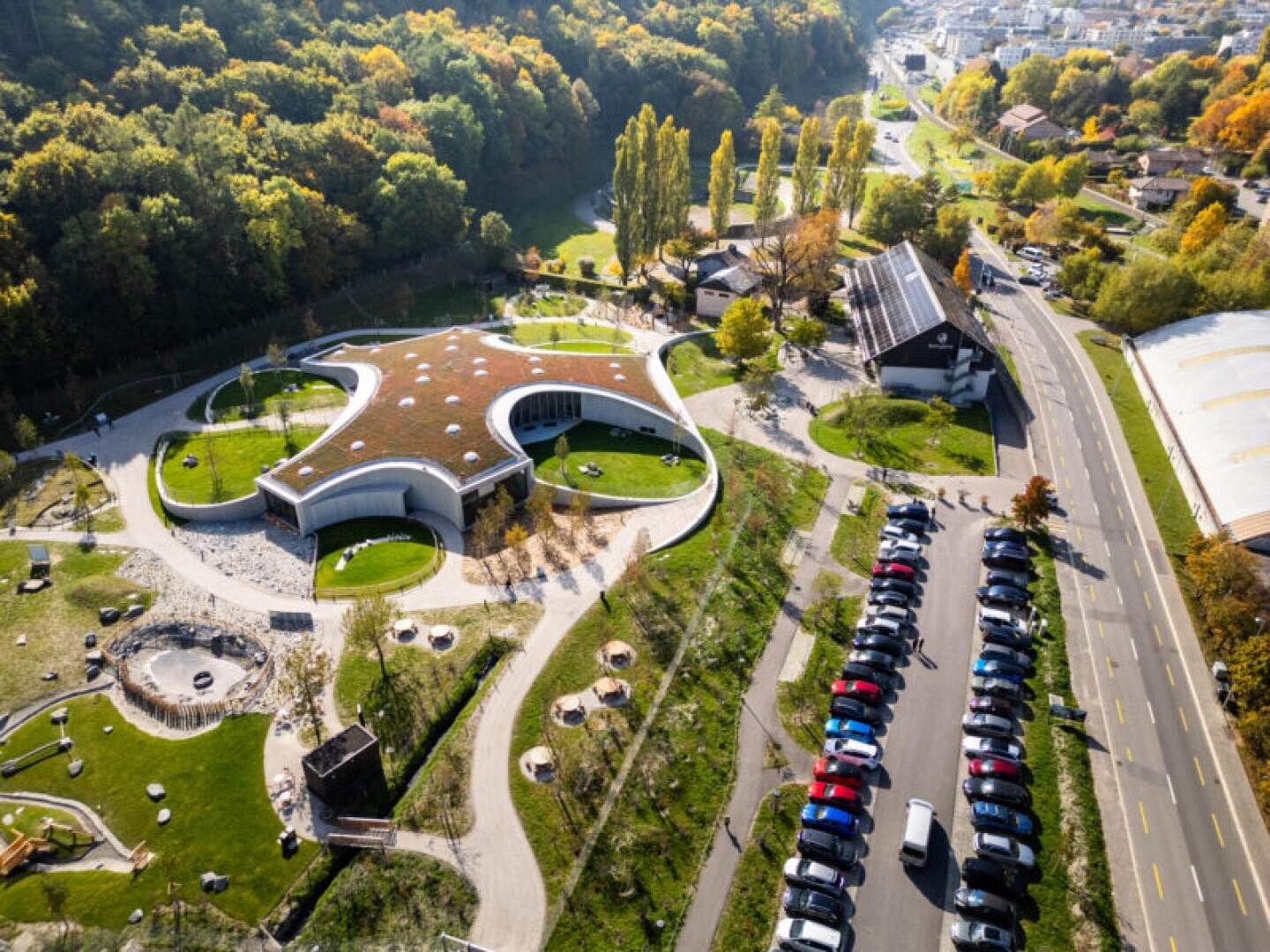 Vue aérienne d'un bâtiment moderne aux lignes courbes, doté d'un toit végétalisé, entouré d'arbres et de pelouses. À proximité, des voitures sont garées le long d'une rue, et d'autres bâtiments sont visibles à l'arrière-plan.