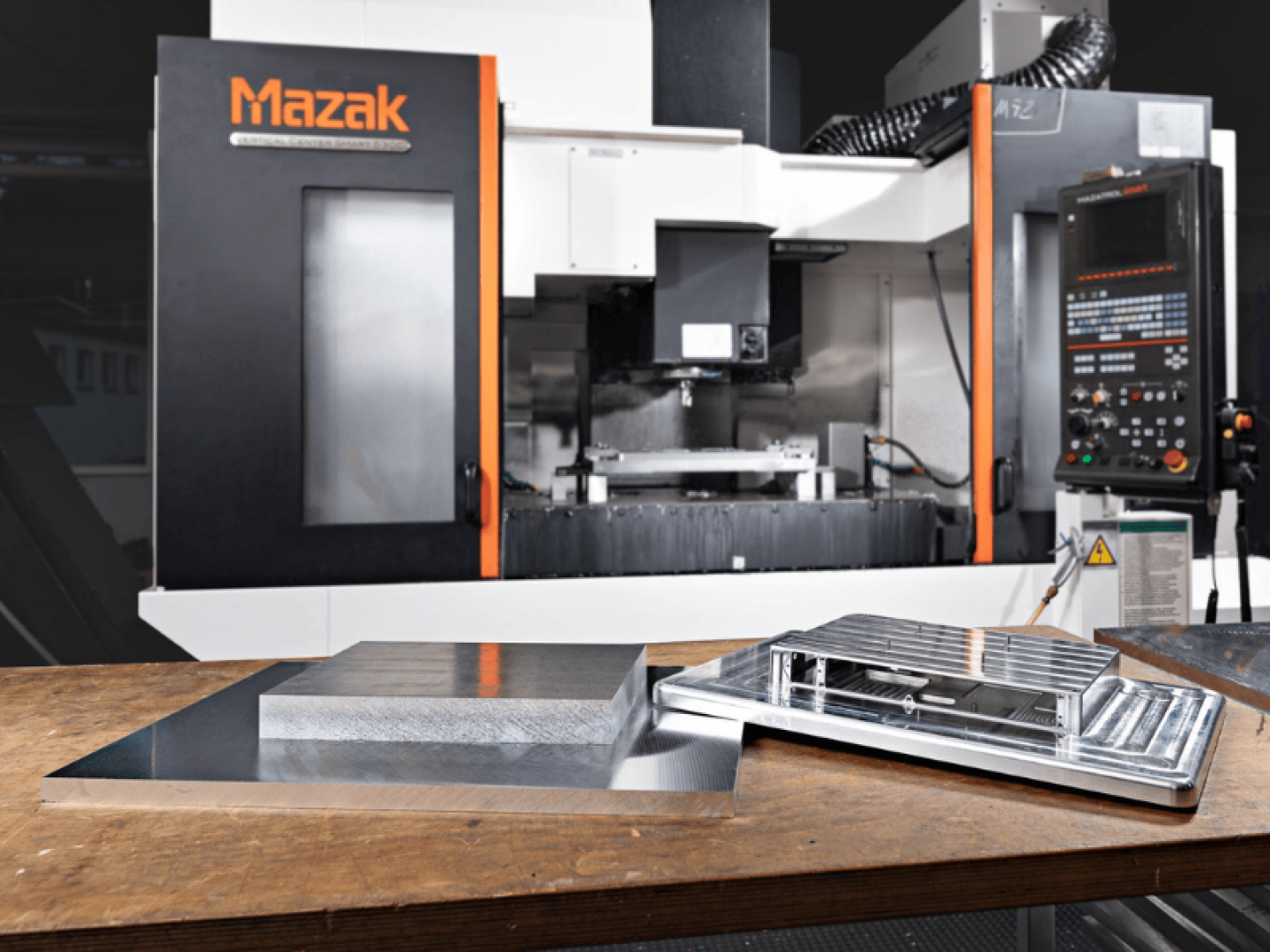 Eine Mazak-CNC-Maschine in einer Werkstatt, im Vordergrund ein Holztisch, auf dem zwei präzisionsgefertigte Metallteile liegen.