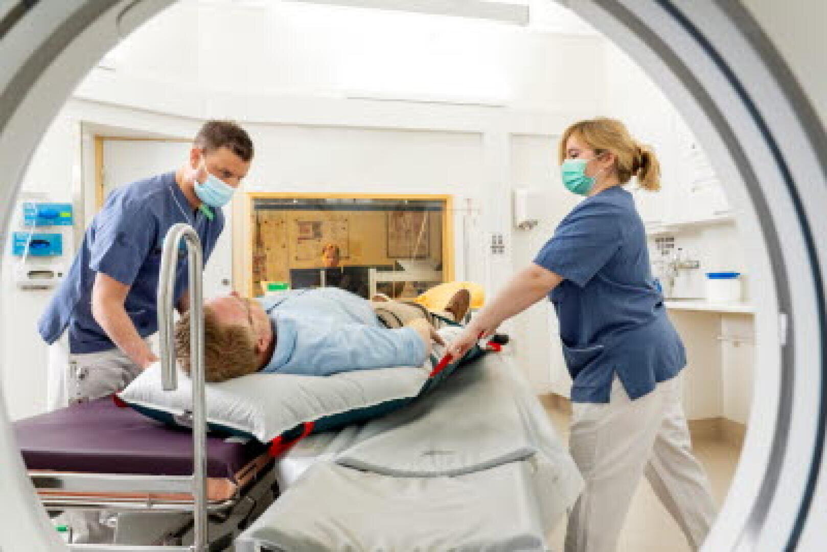 Deux professionnels de santé masqués préparent un patient sur un brancard pour un examen ; la scène est visible à travers l'ouverture circulaire d'un appareil d'imagerie médicale dans une chambre d'hôpital.