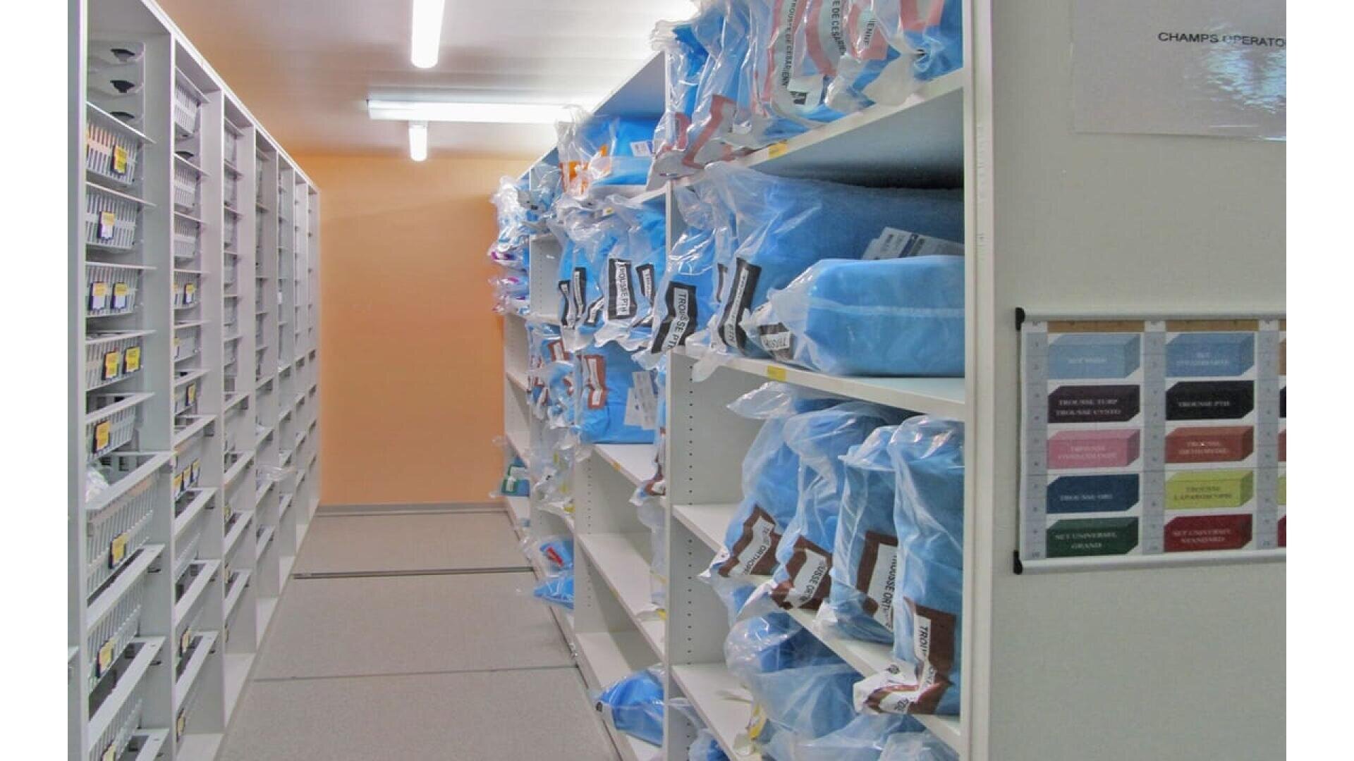 Une salle de stockage avec des étagères contenant des articles emballés en bleu dans des sacs en plastique, chacun étiqueté. La zone est bien éclairée et des tableaux avec un code couleur sont affichés sur le mur de droite.