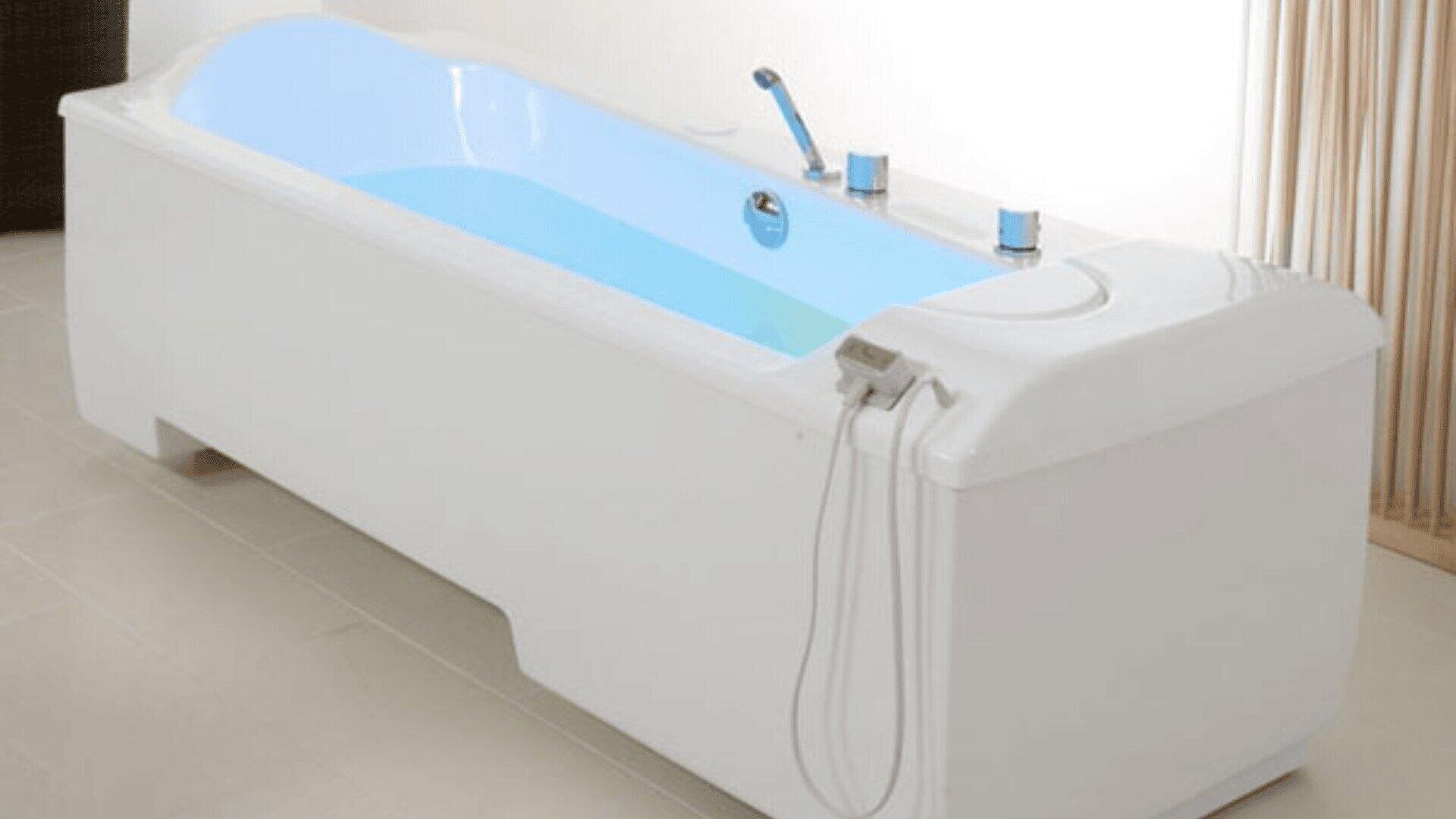 In einem hellen, modernen Badezimmer steht eine weiße Pflegebadewanne, die mit klarem, blauem Wasser gefüllt ist. Die Wanne verfügt über Chromarmaturen und ein Bedienfeld, an dessen einem Ende Duschschläuche befestigt sind.
