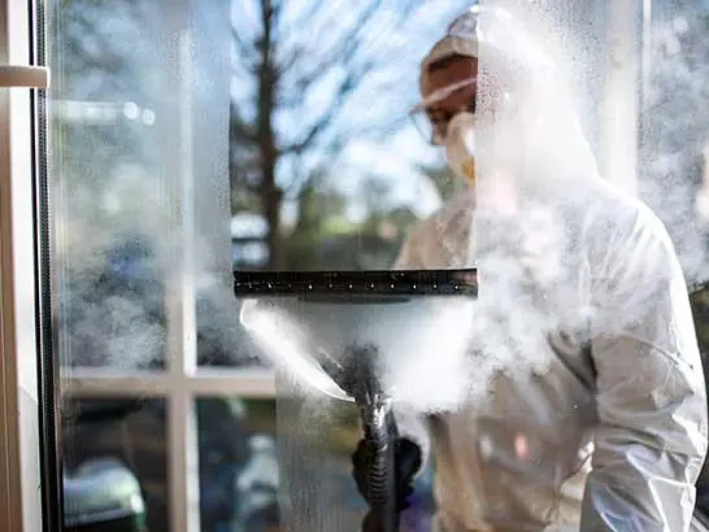 Eine Person in Schutzkleidung und mit Schutzbrille reinigt ein großes Fenster mit einem Abzieher, wodurch Dampf und Wassertropfen auf dem Glas entstehen. Im Hintergrund sind Sonnenlicht und Bäume im Freien zu sehen.