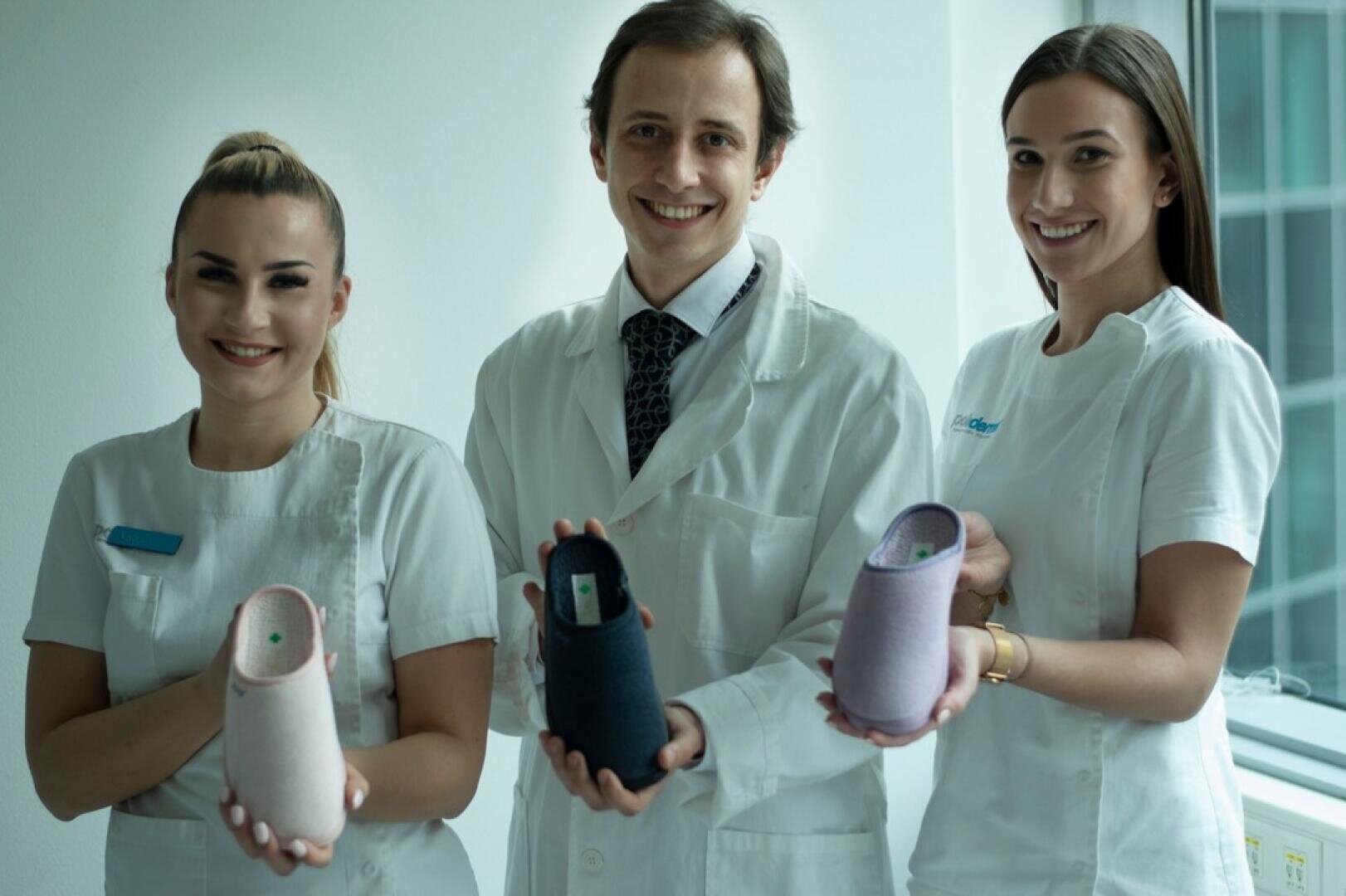Dans une salle de consultation très éclairée, trois personnes souriantes vêtues de blouses blanches de médecin brandissent des chaussons orthopédiques de différentes couleurs.