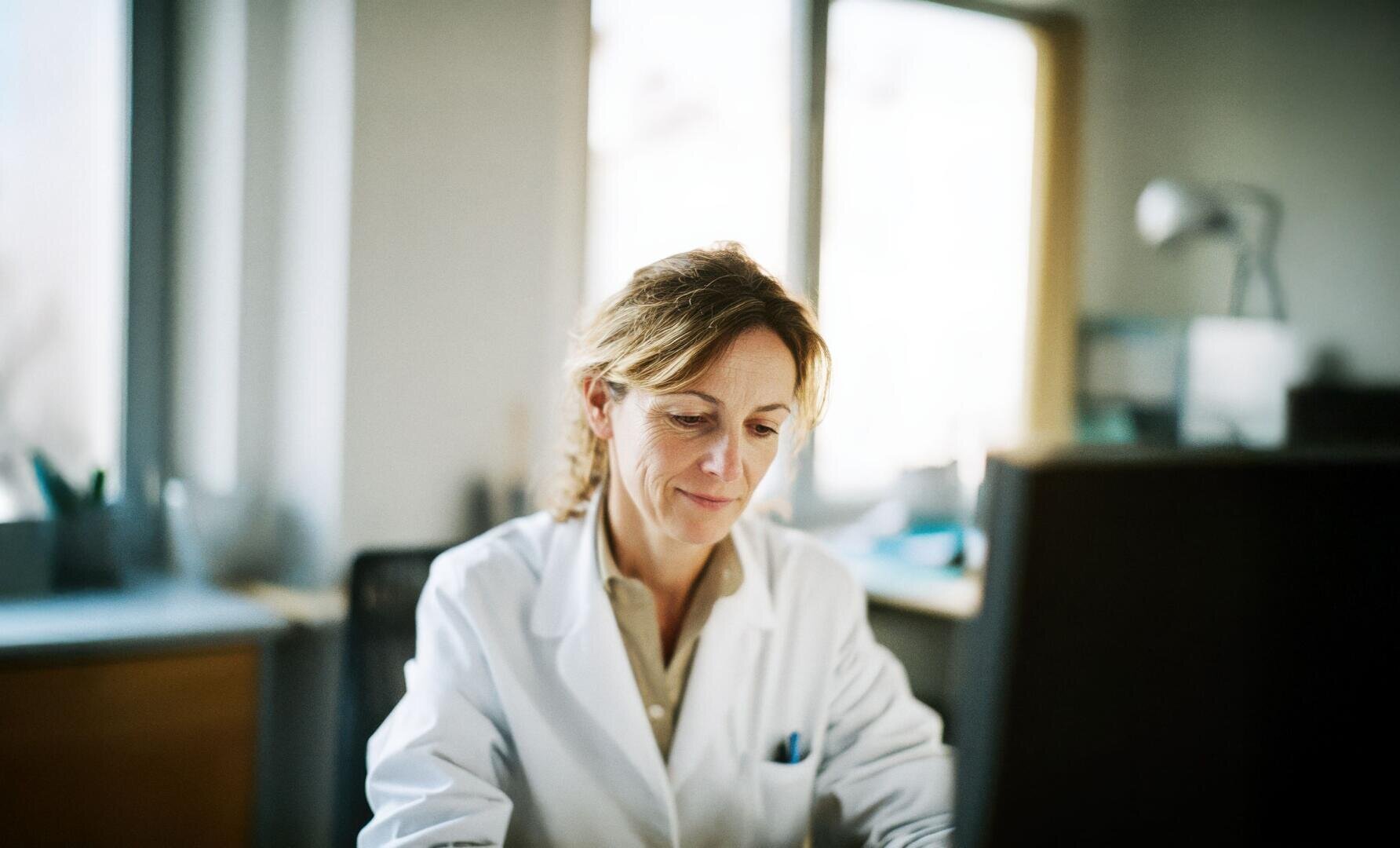 Une femme vêtue d'une blouse blanche de laboratoire est assise à un bureau, les yeux rivés sur l'écran d'un ordinateur, dans un bureau ou un laboratoire bien éclairé.