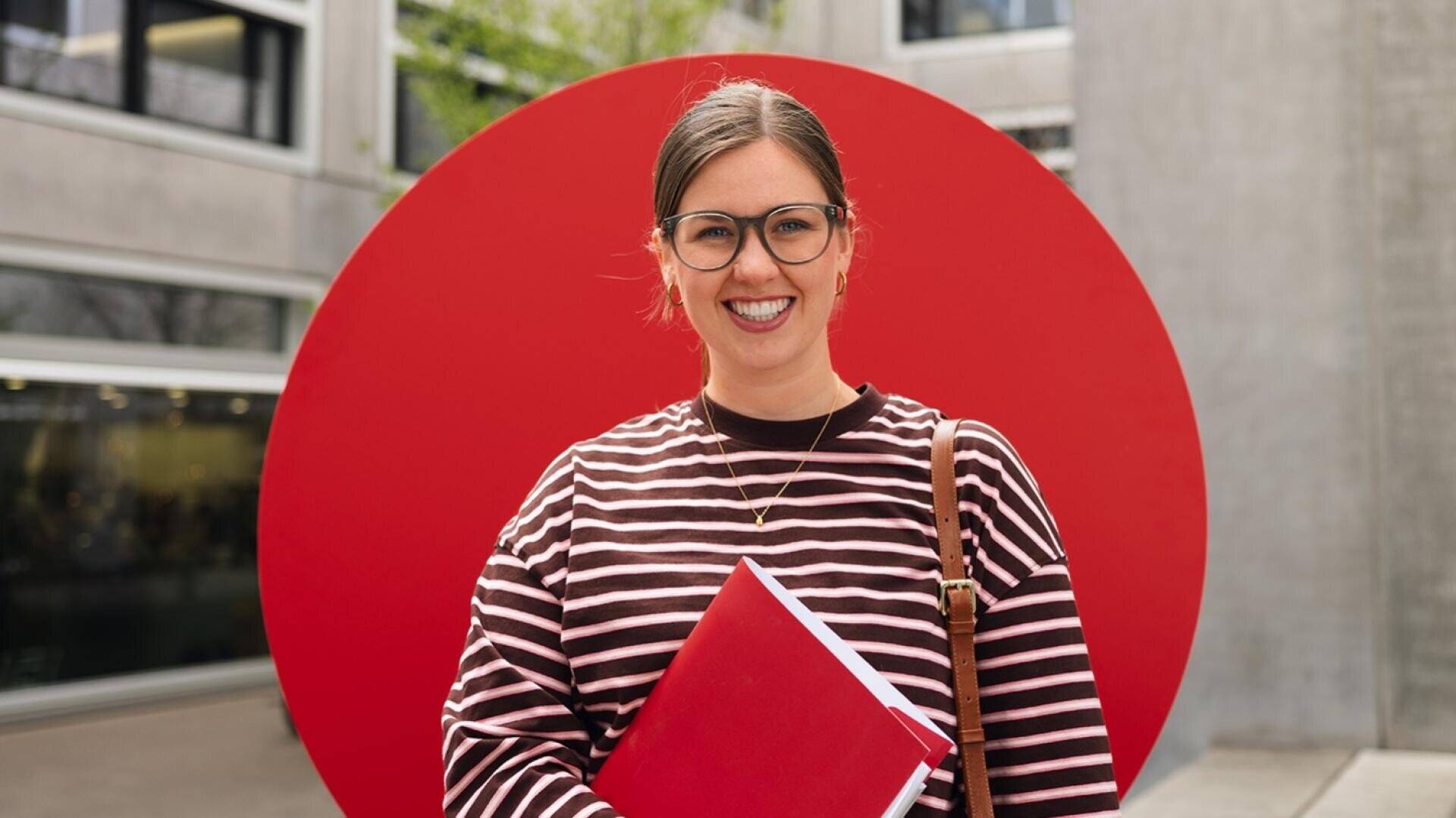 Eine lächelnde Person mit Brille steht vor einem großen roten Kreis vor einem modernen Gebäude und hält eine rote Mappe in der Hand. Sie trägt einen gestreiften Pullover und hat eine braune Umhängetasche dabei.