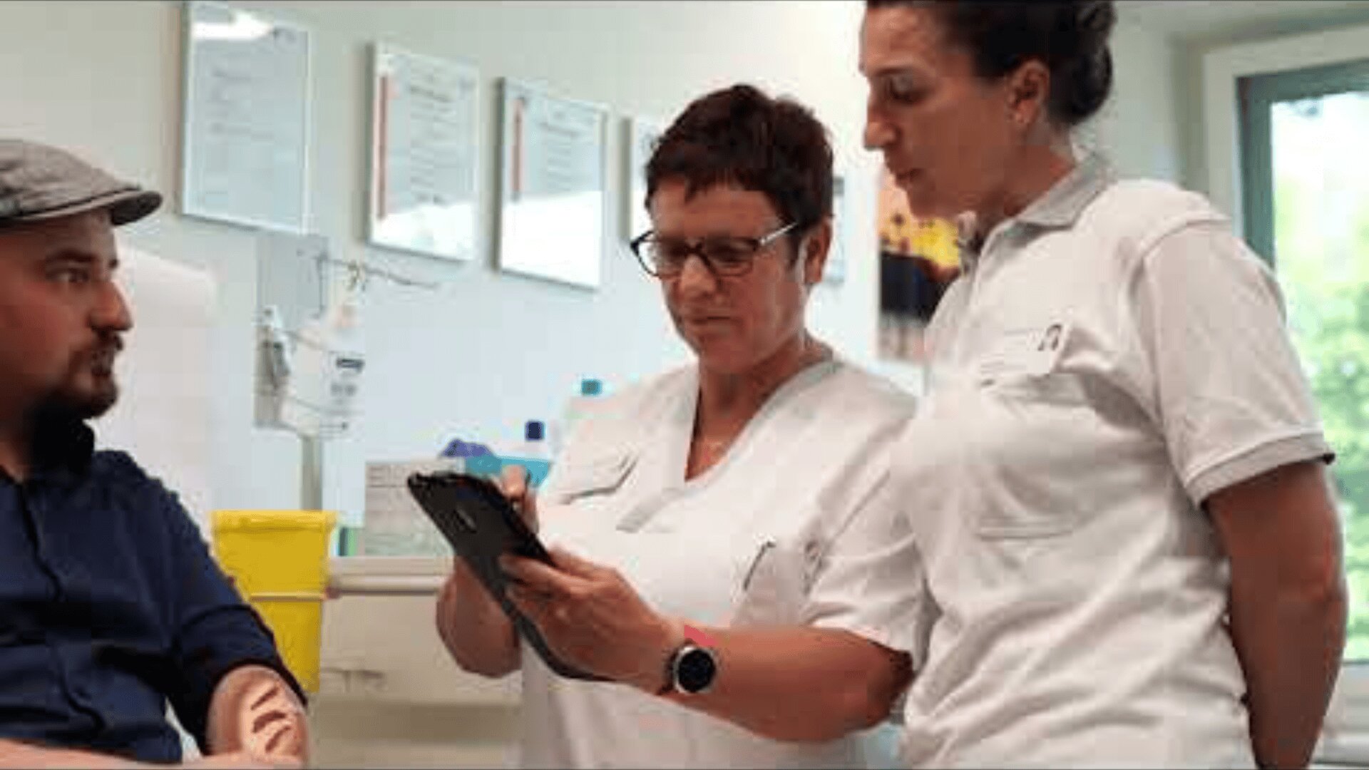 Zwei medizinische Fachkräfte in weißen Uniformen prüfen Informationen auf einem Tablet, während ein sitzender Mann mit einer Mütze zuhört. Die Umgebung scheint eine Arztpraxis oder Klinik zu sein.