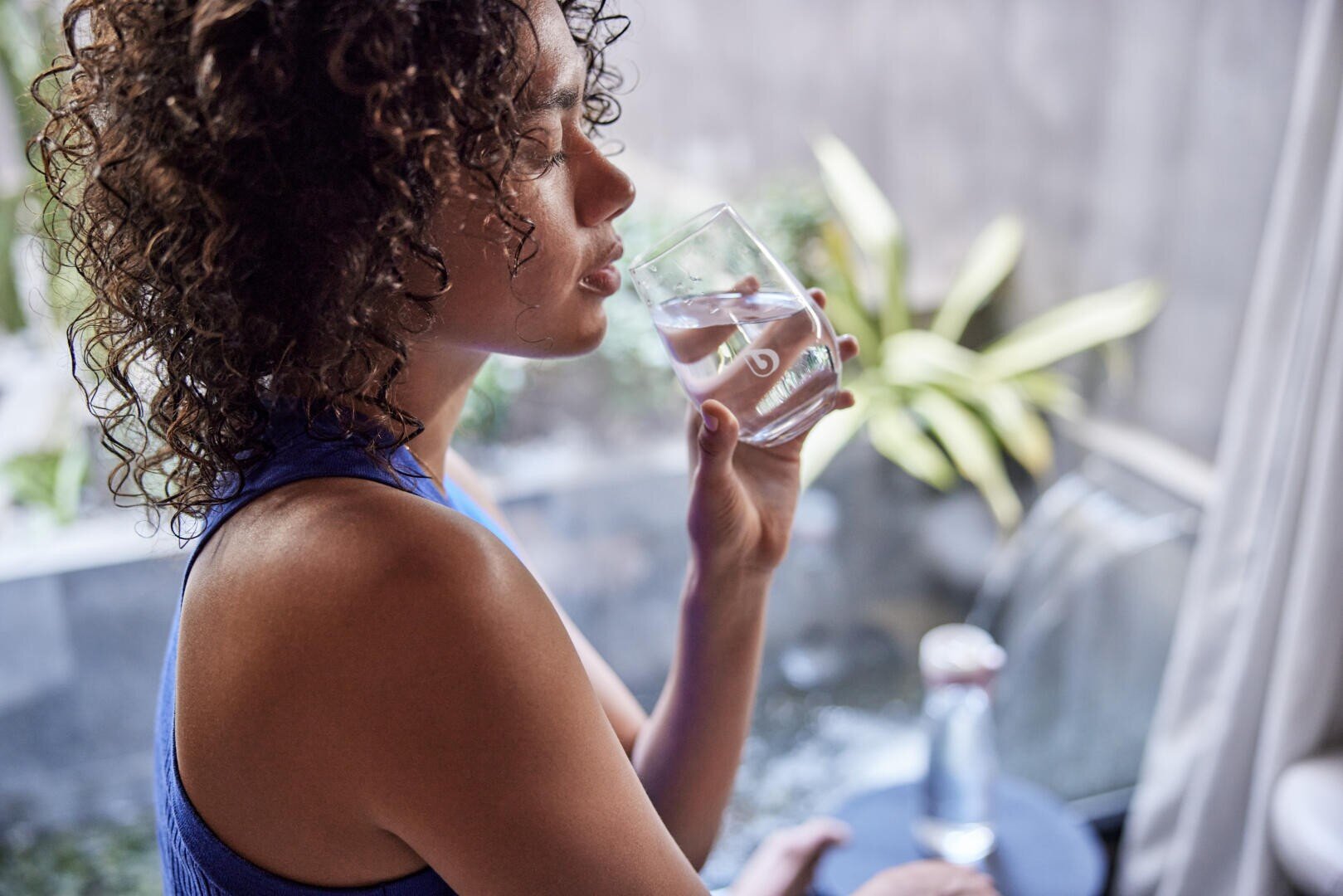Eine Frau mit lockigem Haar in einem blauen Trägertop sitzt drinnen am Fenster und hält ein Glas Wasser an die Lippen; im Hintergrund sind Pflanzen und ein kleines Wasserspiel zu sehen.
