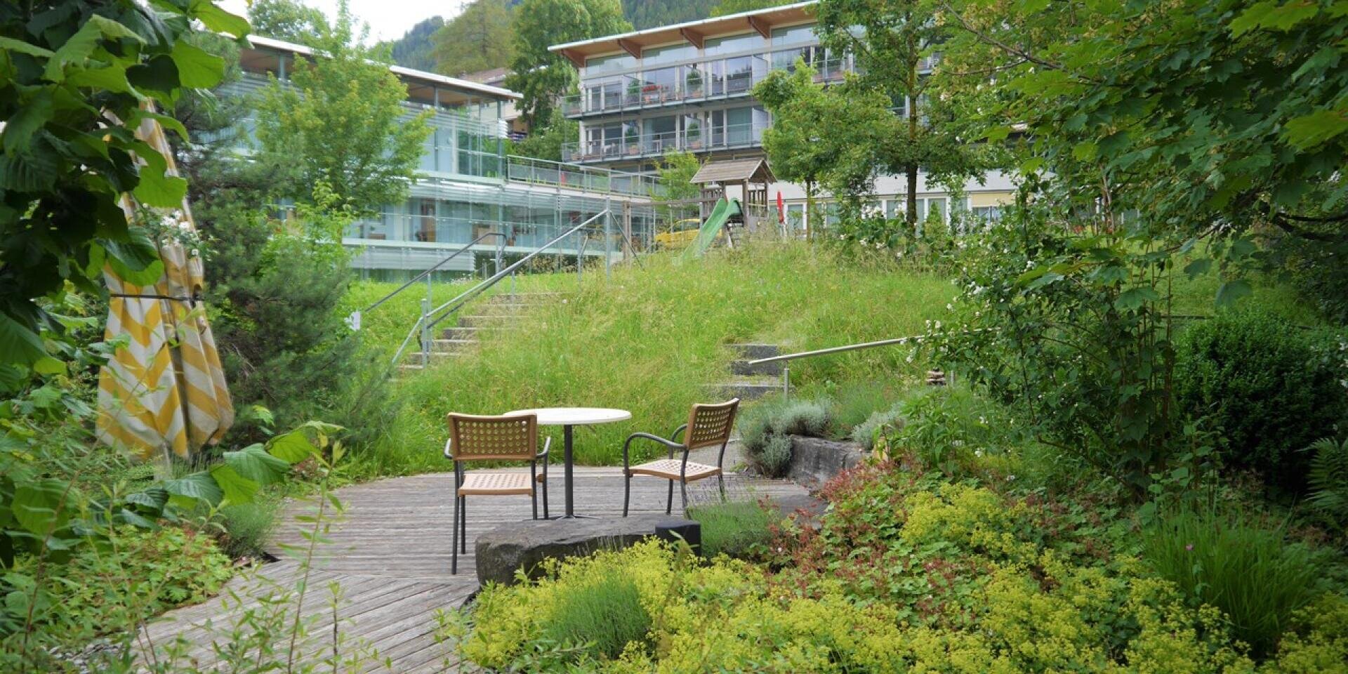 Ein kleiner Tisch und zwei Stühle auf einer Holzterrasse in einem Garten, im Hintergrund ein modernes Gebäude und ein Spielplatz.