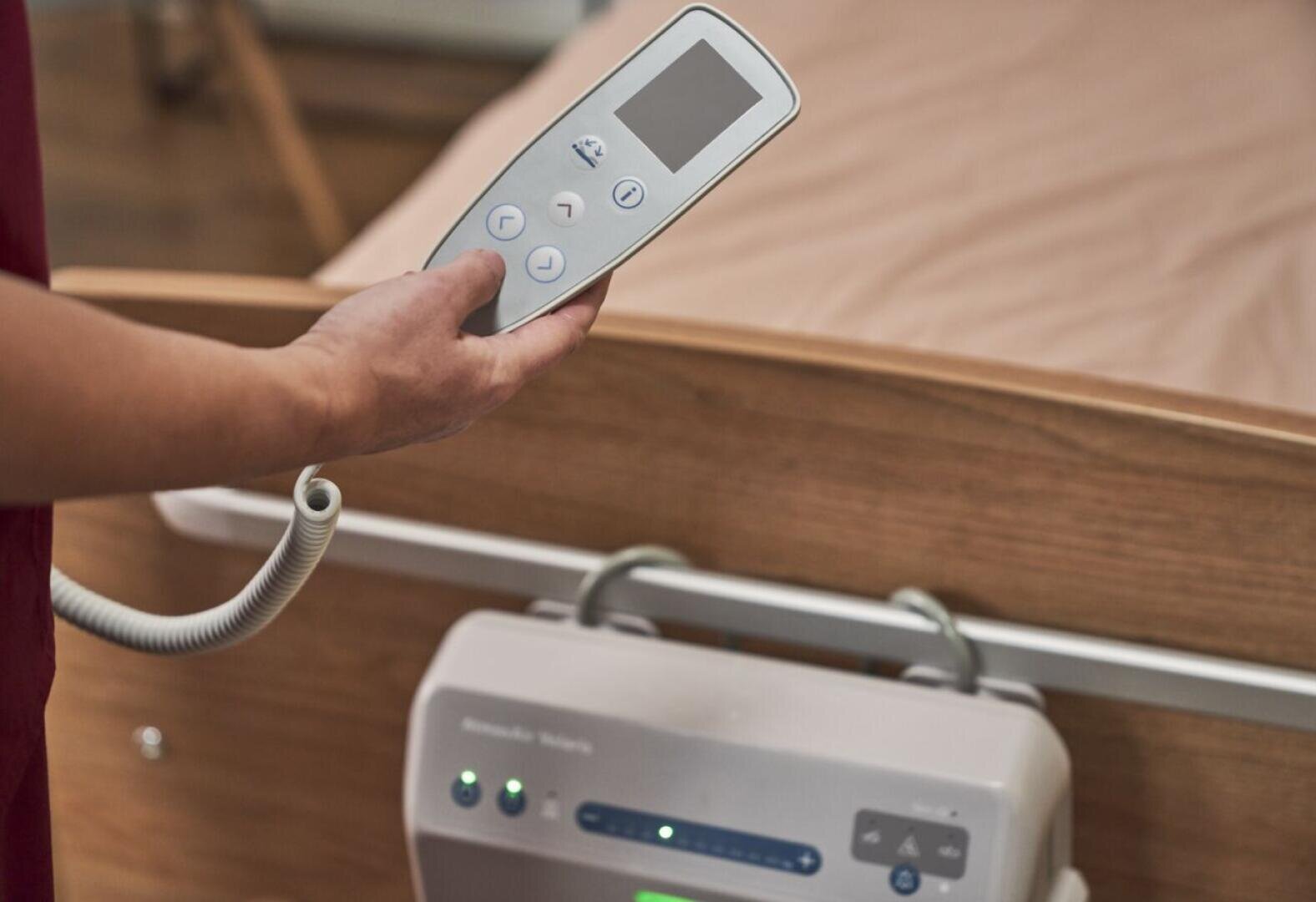 Eine Person hält die Fernbedienung für ein Krankenhausbett in der Nähe eines hölzernen Bettgitters in der Hand. Das Bett ist unbesetzt, und im Vordergrund sind medizinische Geräte zu sehen.