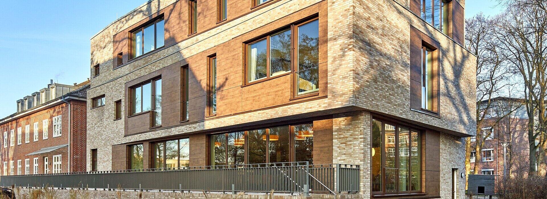 Ein mehrstöckiges Gebäude mit einer Fassade aus hellbeigen Ziegeln und braunen Verkleidungsplatten, großen Fenstern und einer Terrasse im Erdgeschoss, die von einem Metallgeländer umgeben ist.
