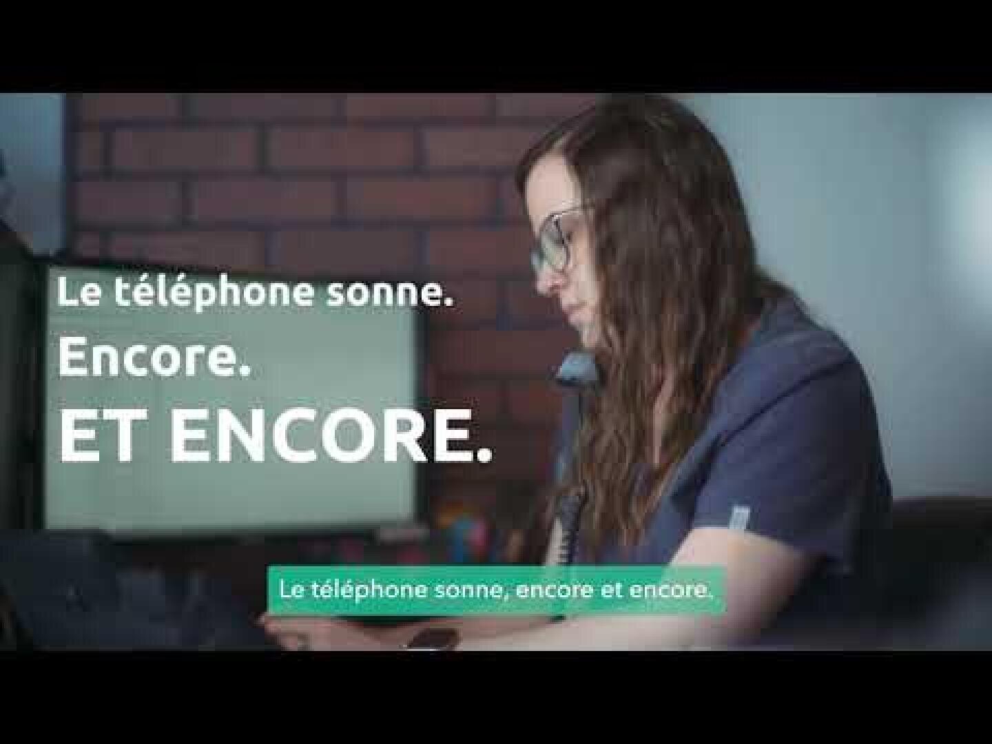 Une femme aux longs cheveux châtains et portant des lunettes est au téléphone, assise à un bureau. Le texte en français sur l'image indique : « Le téléphone sonne. Encore. ET ENCORE. » Une bande verte indique également : « Le téléphone sonne, encore et encore. ».