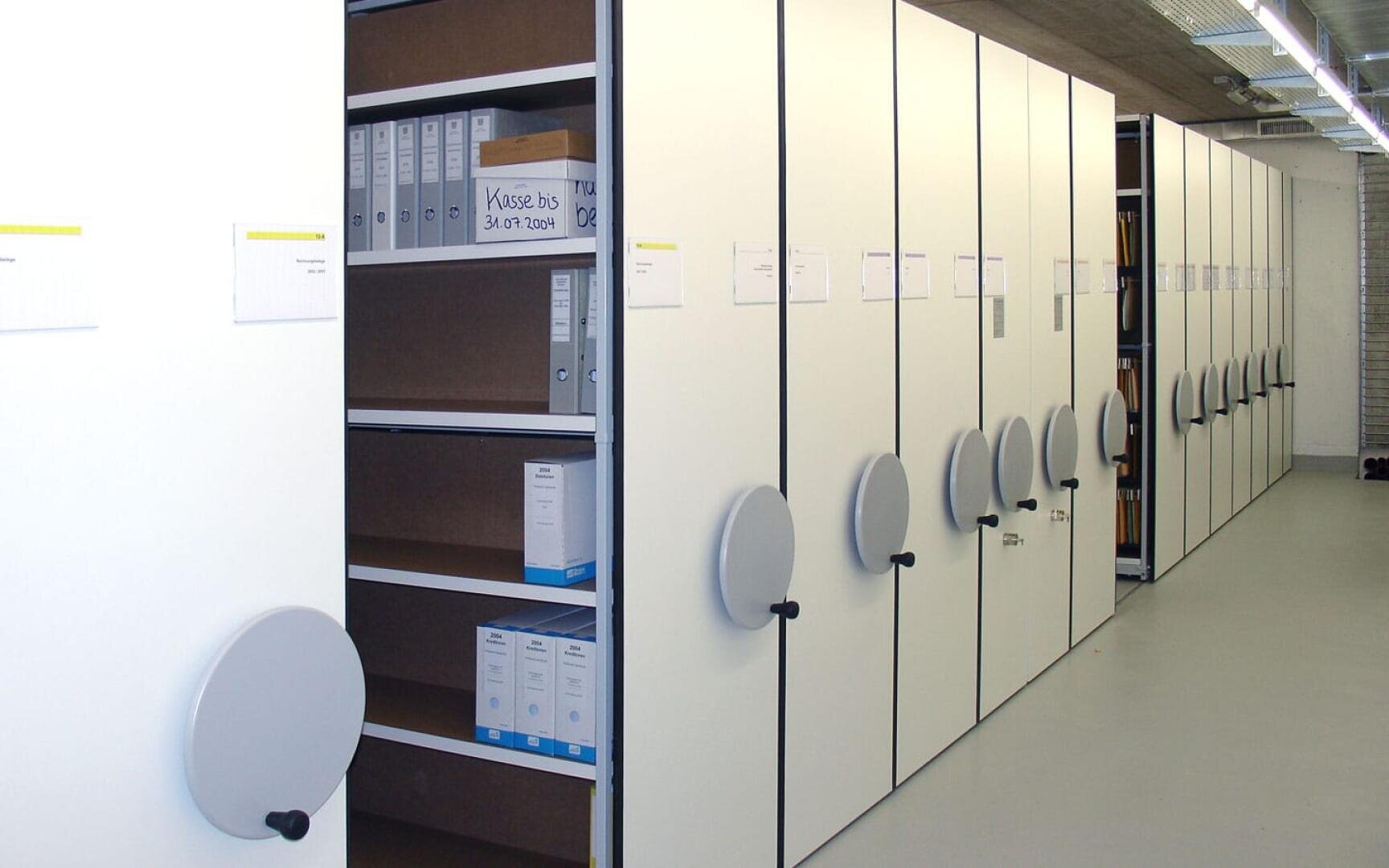 Eine Reihe weißer, mobiler Regale mit großen runden Griffen in einem Büro oder Archiv, von denen einige teilweise geöffnet sind, um Regale mit beschrifteten Ordnern und Akten freizugeben. Der Boden ist hellgrau und die Decke ist im industriellen Stil gehalten.