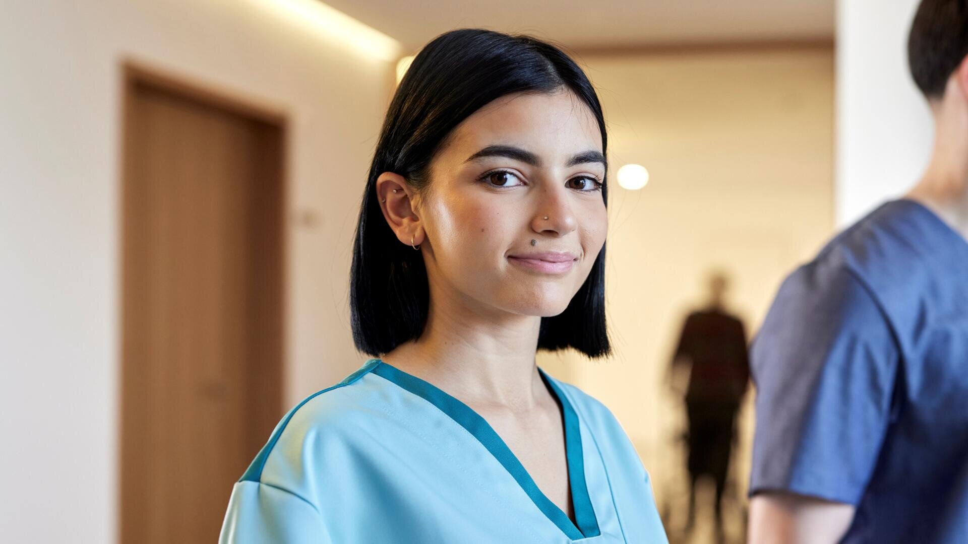 Une jeune femme aux cheveux noirs courts, vêtue d'une tenue médicale, sourit à la caméra dans un couloir d'hôpital.