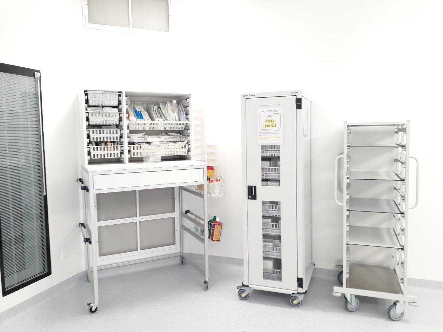 Une salle de fournitures médicales claire et propre avec trois unités de stockage métalliques sur roues : une avec des fournitures médicales organisées, une armoire haute avec des sections transparentes et une étagère avec des plateaux vides, le tout sur des murs blancs et un sol gris.