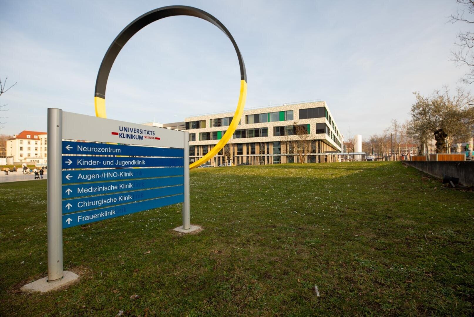 Sur une pelouse devant un bâtiment hospitalier moderne, un panneau indicateur bleu et blanc ; une grande sculpture ronde est visible à l'arrière-plan. Le panneau indique « Clinique universitaire » et énumère les services cliniques.