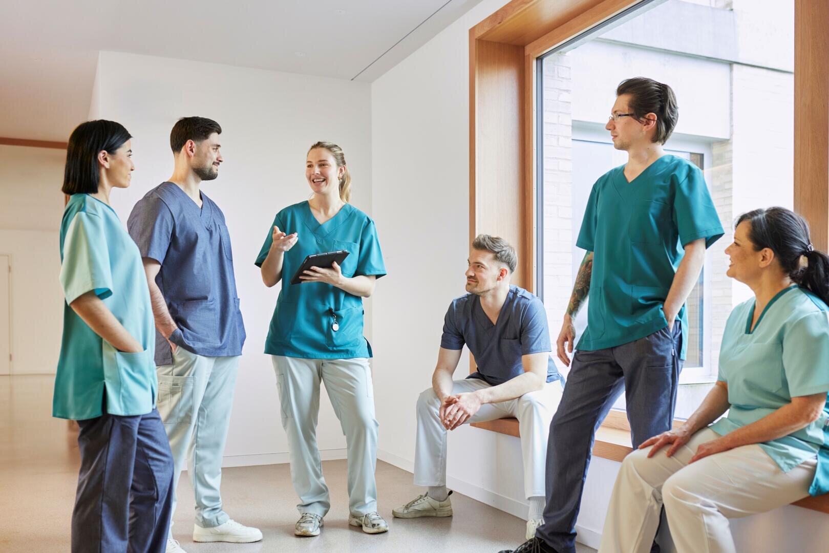 Six professionnels de santé en tenue chirurgicale discutent et s'échangent des sourires dans un couloir lumineux ; certains sont debout, tandis que d'autres sont assis près d'une grande fenêtre.
