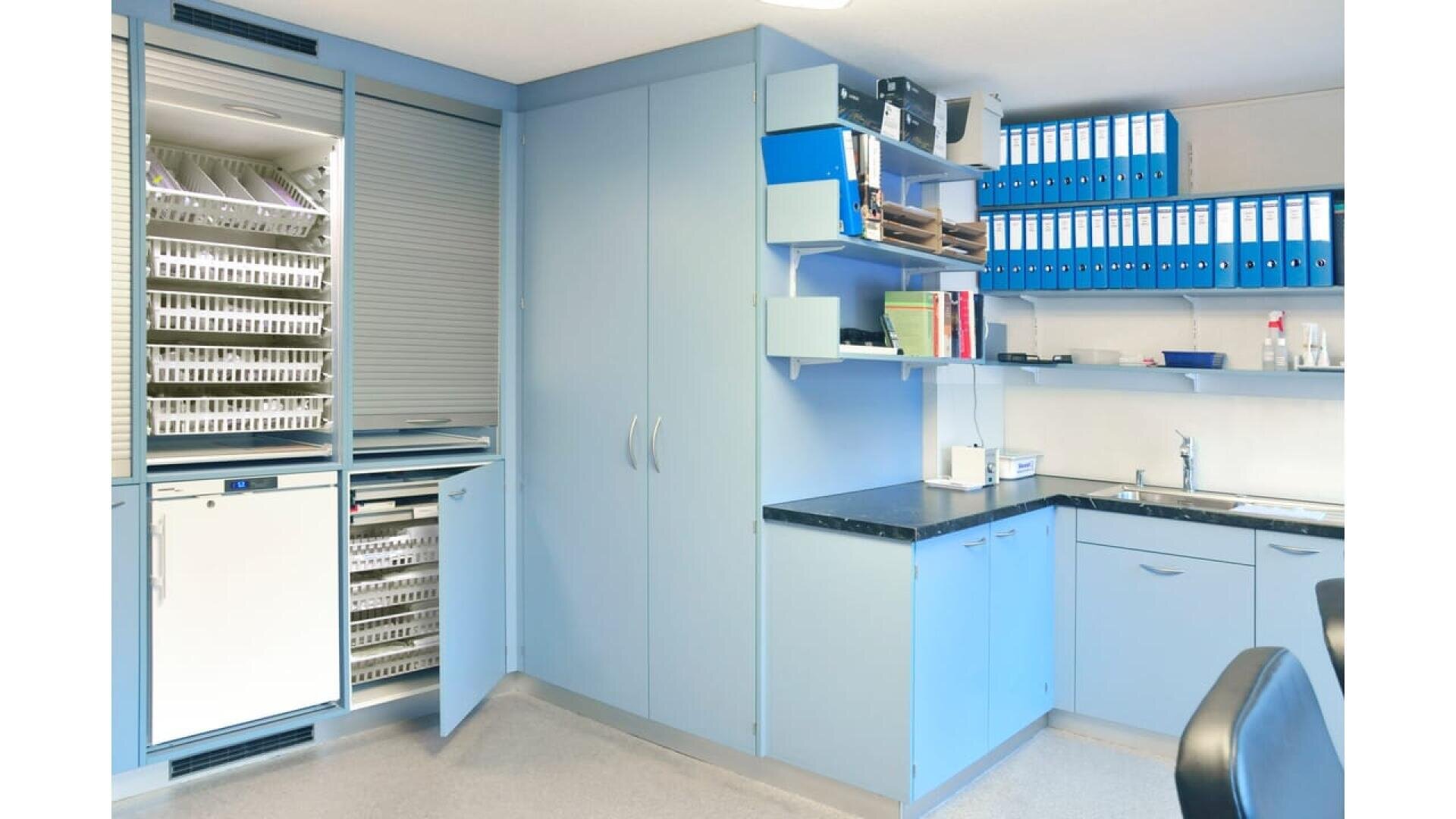 Un cabinet médical ou dentaire propre et moderne avec des armoires bleues, des étagères contenant des dossiers et des classeurs, un évier et des espaces de rangement organisés. L'espace semble lumineux et bien organisé.