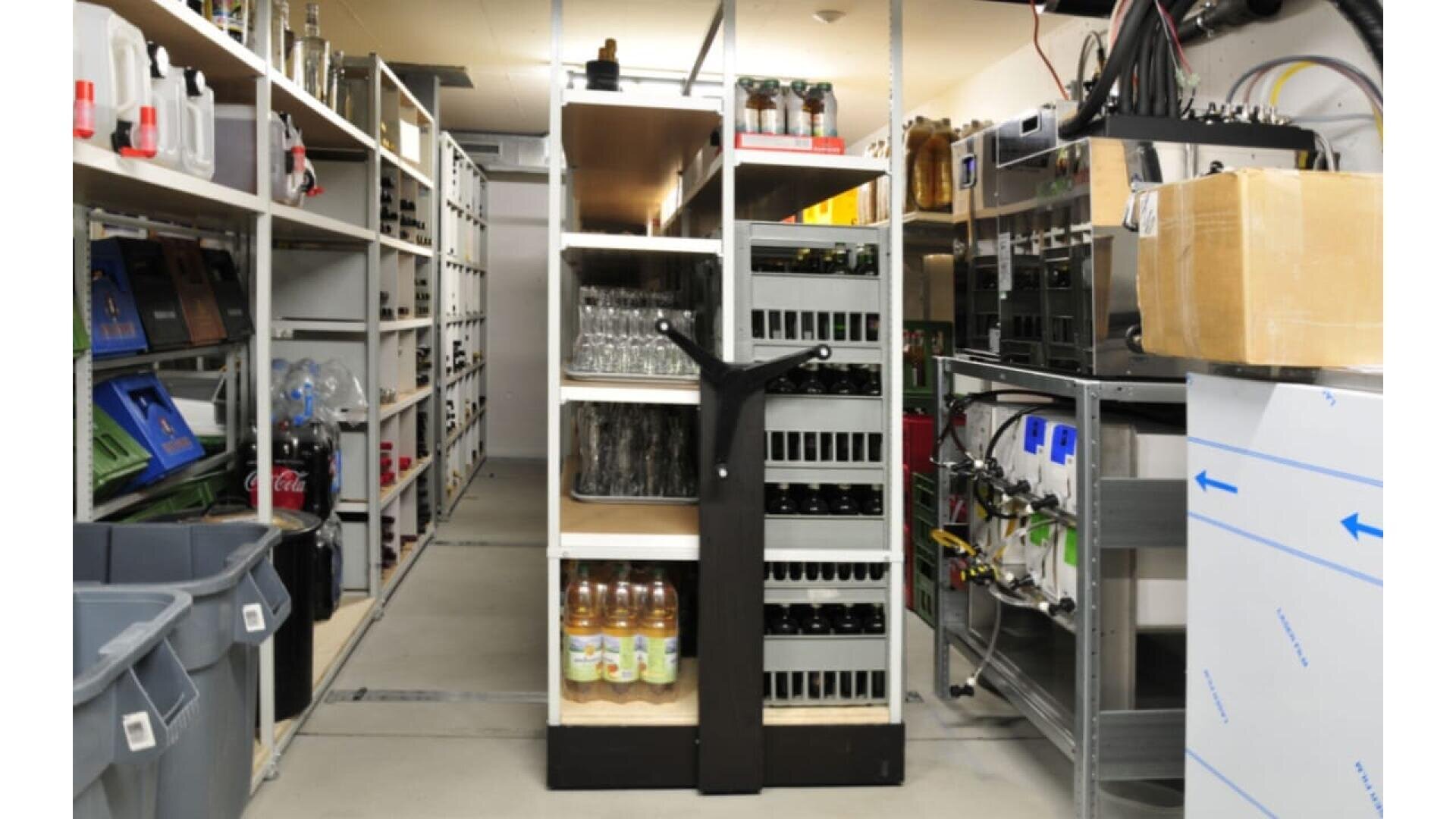 Une salle de stockage bien organisée avec des étagères contenant des bouteilles, des boissons, des fournitures de cuisine et des boîtes. Il y a des bacs sur la gauche, de la verrerie et des bouteilles de boissons au centre, et divers équipements et fournitures sur la droite.