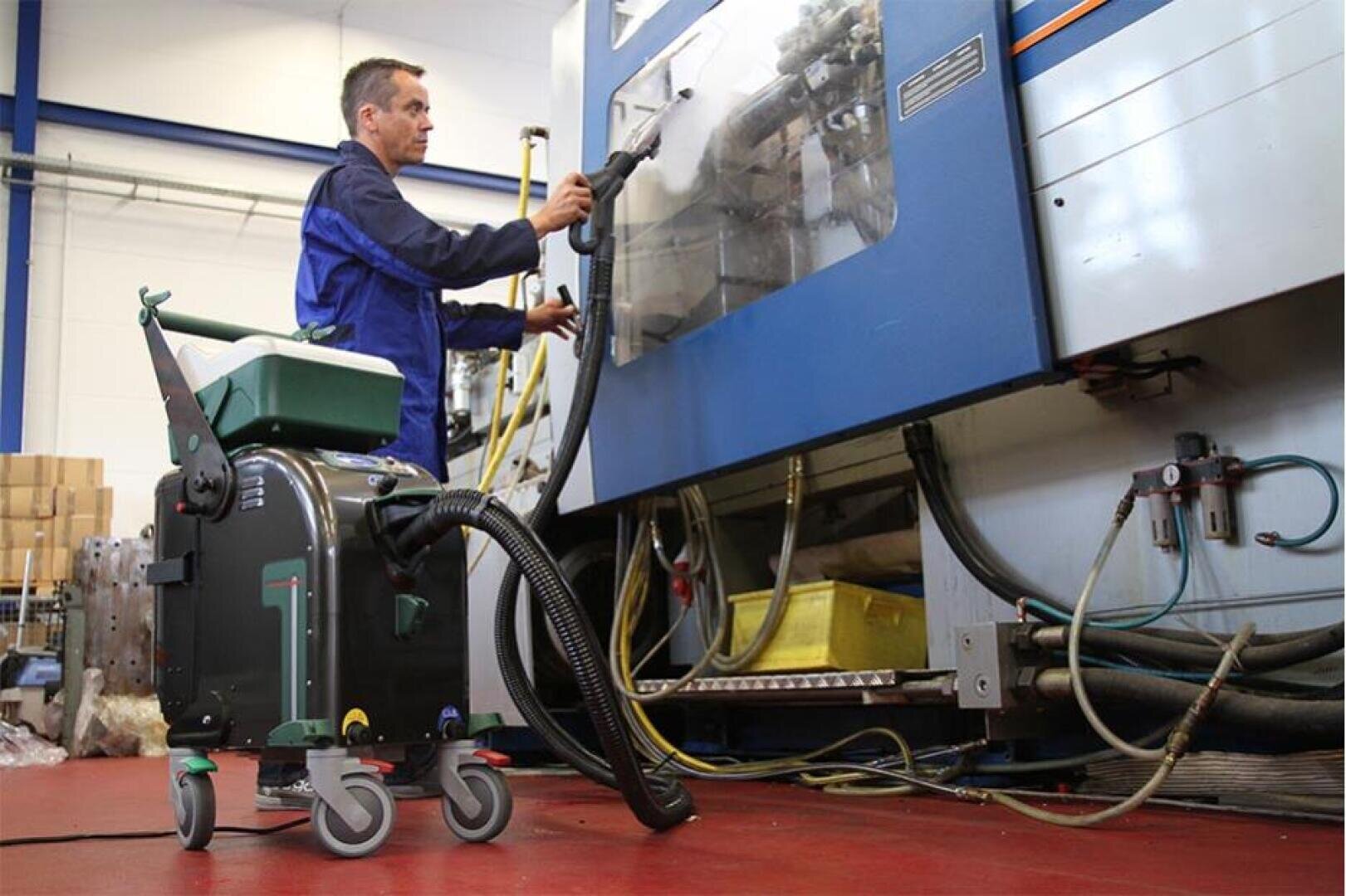 Eine Person in Arbeitskleidung reinigt mit einer Saugmaschine das Innere einer großen blauen Industriemaschine in einer Fabrik. Der Boden ist rot, und es sind verschiedene Rohre und Schläuche zu sehen.