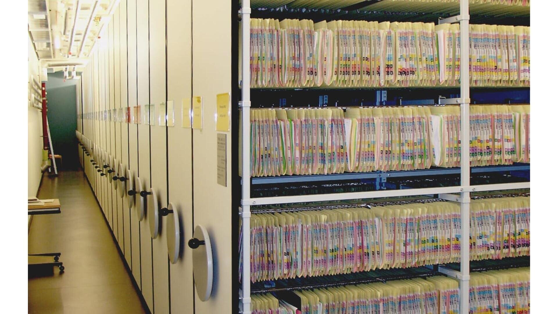 Des rangées de rayonnages mobiles remplis de dossiers organisés et codés par couleur, probablement dans une salle de stockage de documents ou d'archives. Les étagères sont densément remplies, avec des poignées permettant de déplacer les sections pour accéder aux dossiers.