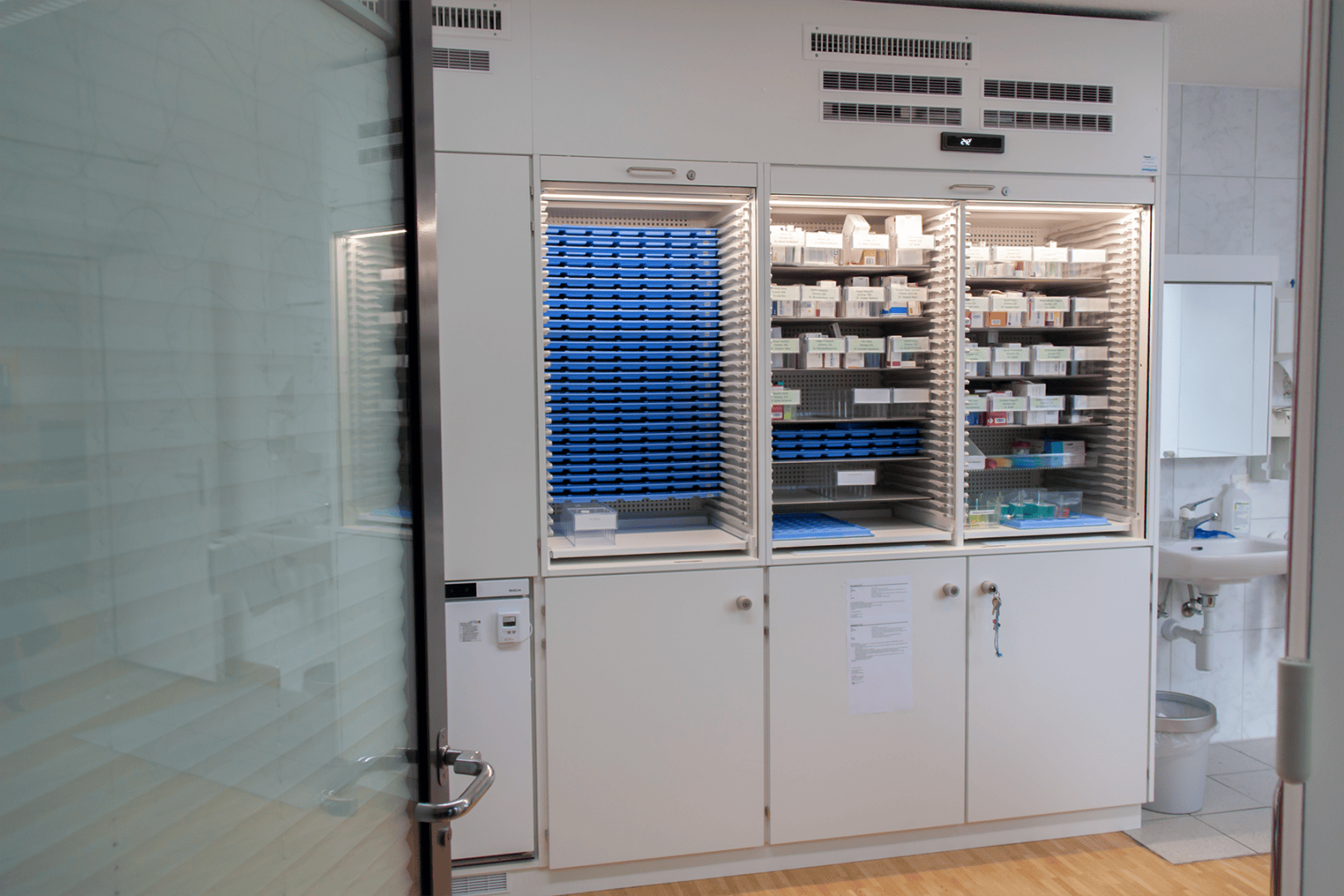 Ein Apothekenschrank mit offenen Türen gibt den Blick auf übersichtliche Regale frei, die mit verschiedenen Medikamentenschachteln, Tablettenbehältern und blauen Tabletts gefüllt sind; in der Nähe sind eine Glastür und ein Waschbecken zu sehen.