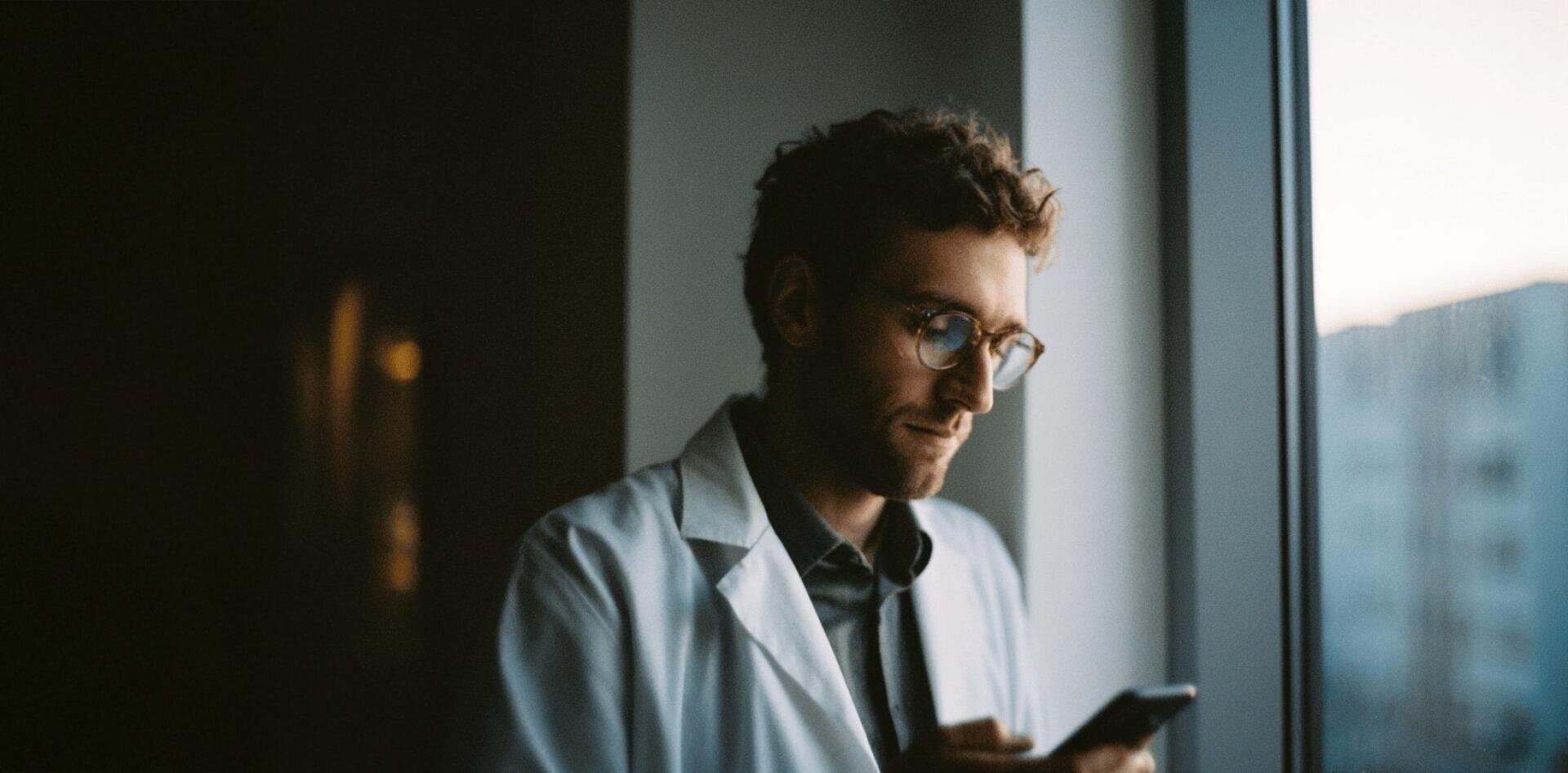 Une personne vêtue d'une blouse blanche de laboratoire et portant des lunettes se tient près de la fenêtre, le regard baissé sur un smartphone. L'arrière-plan est légèrement éclairé ; on aperçoit des bâtiments flous à l'extérieur.