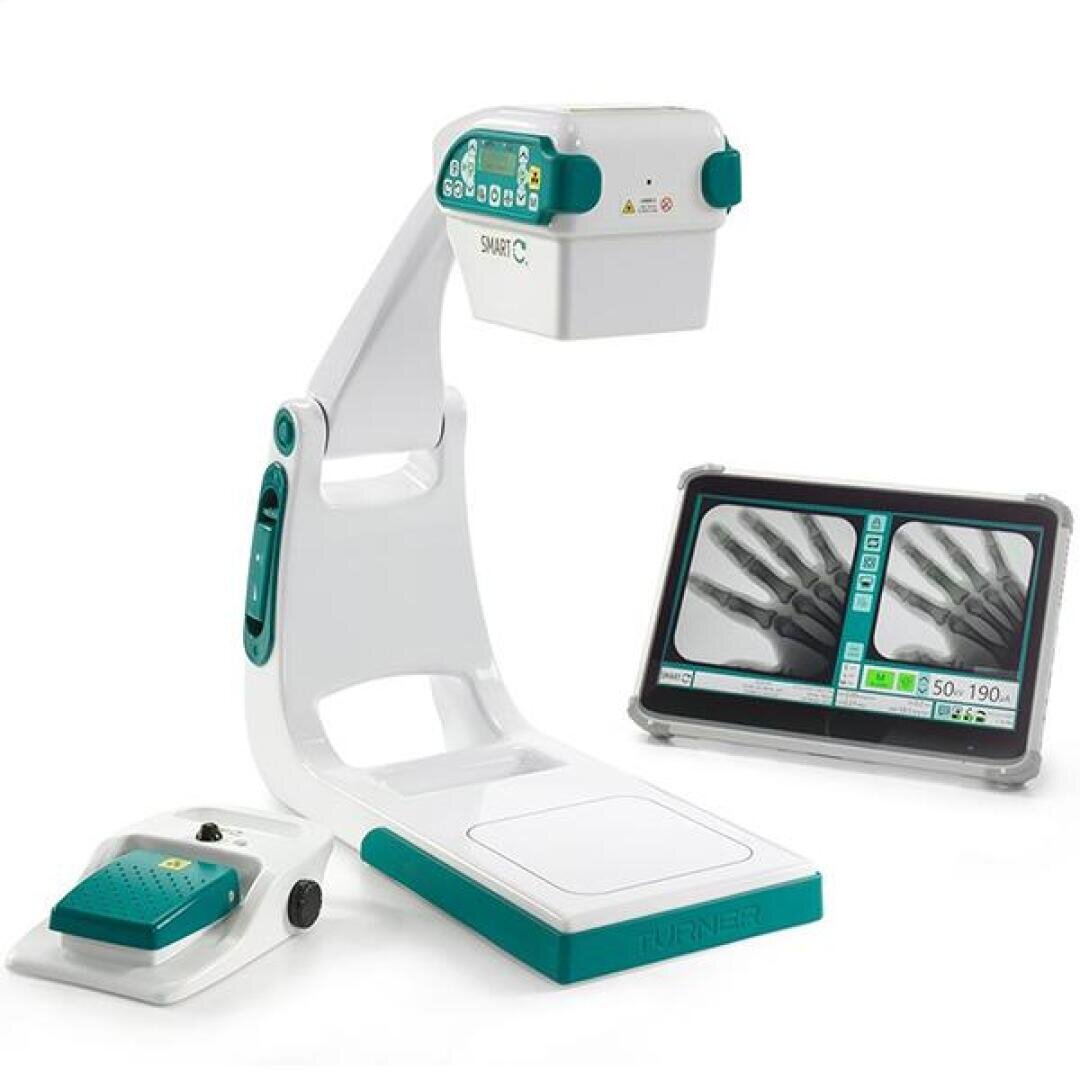 Un appareil d'imagerie médicale compact équipé d'un écran intégré sur lequel s'affichent des radiographies des doigts, ainsi que d'une pédale de commande située sur le côté ; le tout dans des tons de blanc et de bleu-vert.