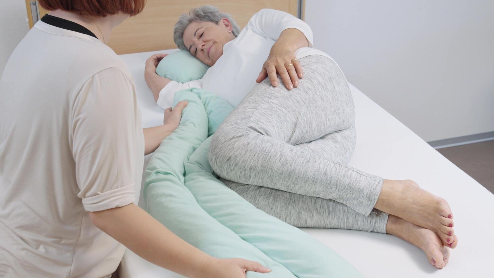 Une personne âgée est allongée sur le côté dans son lit, serrant contre elle un grand oreiller de soutien, tandis qu'une autre personne est assise à ses côtés, lui apportant son aide ou son réconfort.