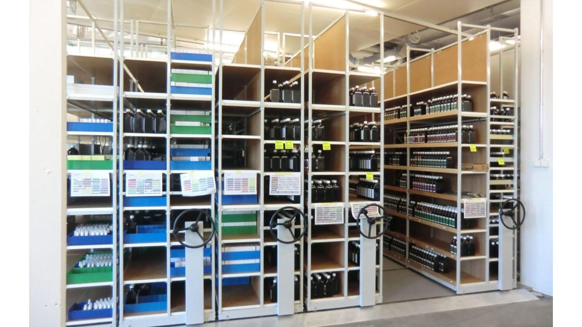 Des étagères remplies de rangées de bouteilles sont organisées dans une salle de stockage. Les rayonnages sont équipés de grandes manivelles pour un accès glissant, et des étiquettes et des panneaux de couleur sont visibles sur les rayonnages et les bouteilles.