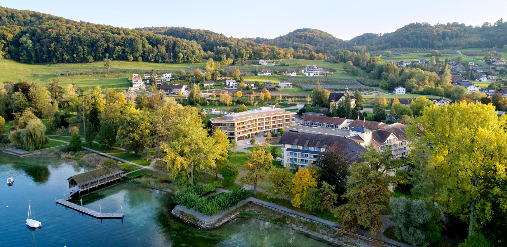 Vue aérienne de bâtiments situés au bord d'un lac, entourés d'arbres, de collines et de maisons isolées, avec une jetée en bois qui s'avance dans l'eau limpide sous un ciel bleu.