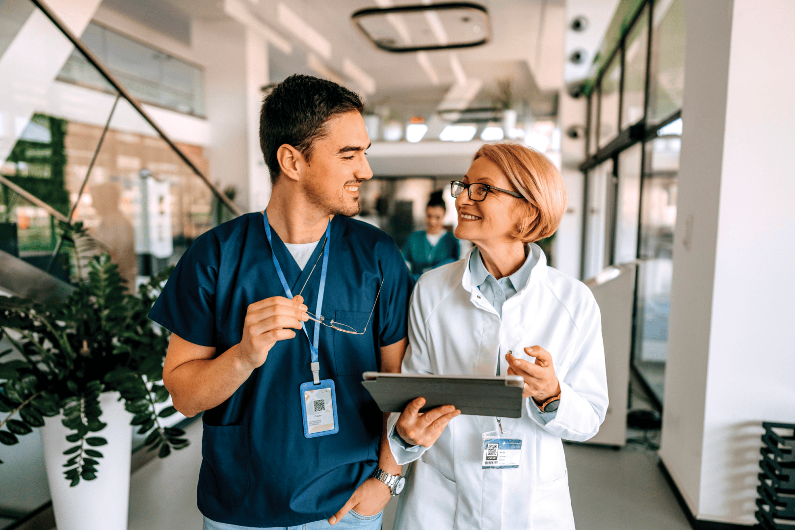 Zwei lächelnde medizinische Fachkräfte, eine in blauer OP-Kleidung und die andere in einem weißen Kittel, gehen gemeinsam durch einen Krankenhausflur, unterhalten sich und schauen auf ein Tablet.