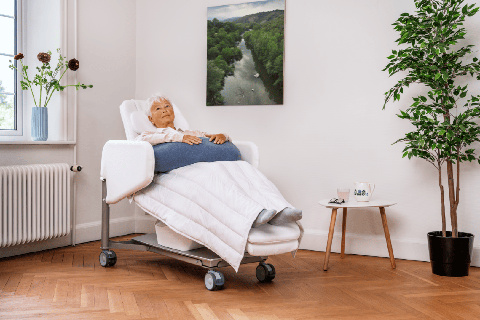 Une femme âgée s'installe confortablement dans un fauteuil inclinable, enveloppée dans une couverture, dans une pièce lumineuse décorée de plantes, de fleurs et d'un tableau représentant un paysage accroché au mur.