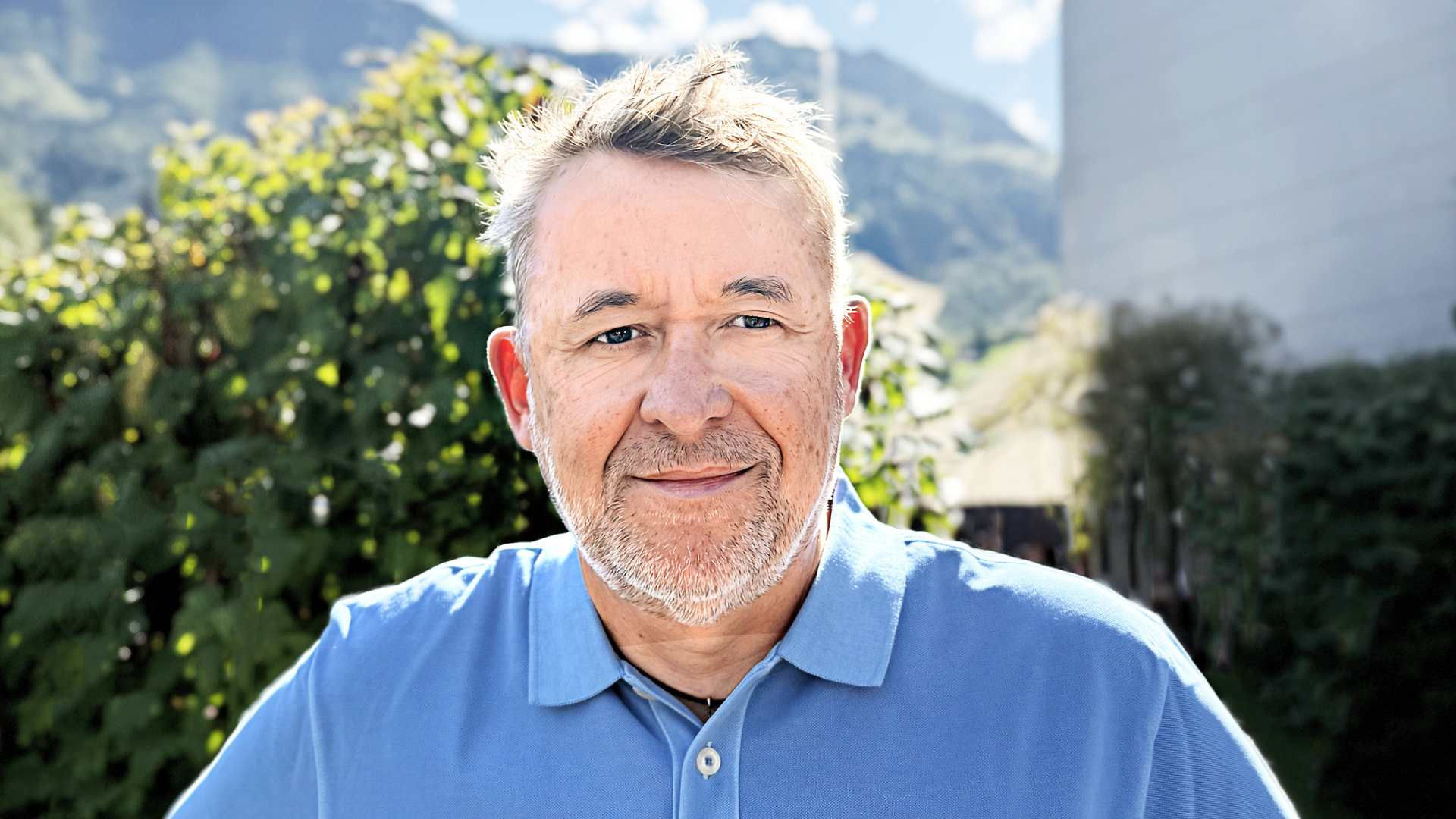 Ein Mann mittleren Alters mit kurzen grauen Haaren und Bart, der ein blaues Poloshirt trägt, steht im Freien im Sonnenlicht, im Hintergrund sind grünes Laub und Berge zu sehen.