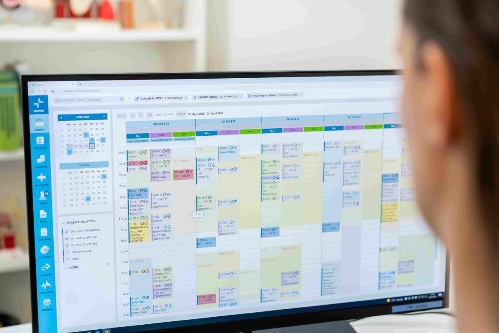 Eine Person schaut auf einen Computermonitor, auf dem ein detaillierter, farbenfroher digitaler Kalender oder eine Planungssoftware angezeigt wird, in der verschiedene Termine und Ereignisse in Spalten angeordnet sind.