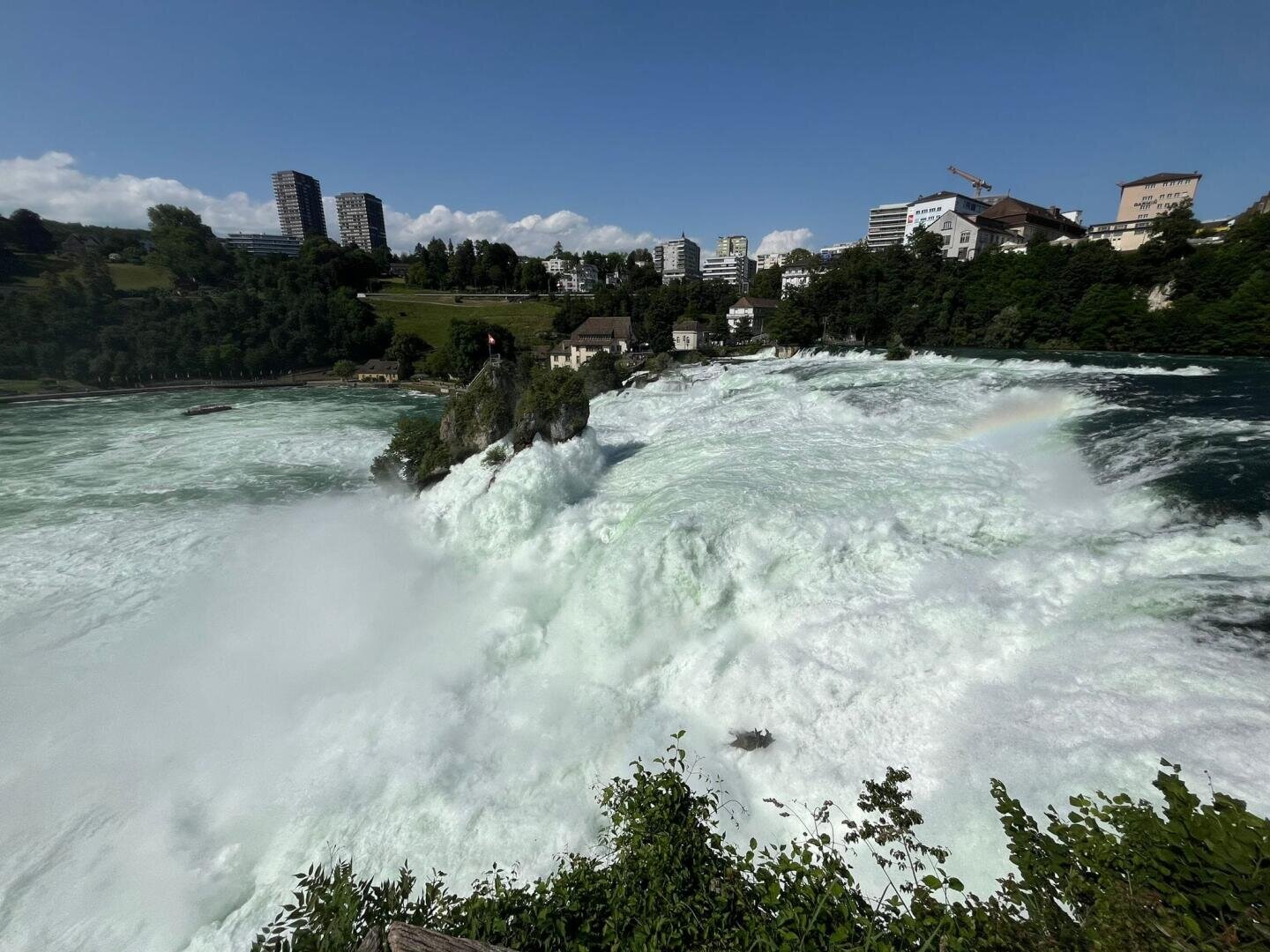 Ein mächtiger Wasserfall fließt mit weißem Nebel und einem schwachen Regenbogen unter einem klaren blauen Himmel, umgeben von Grün und städtischen Gebäuden im Hintergrund.