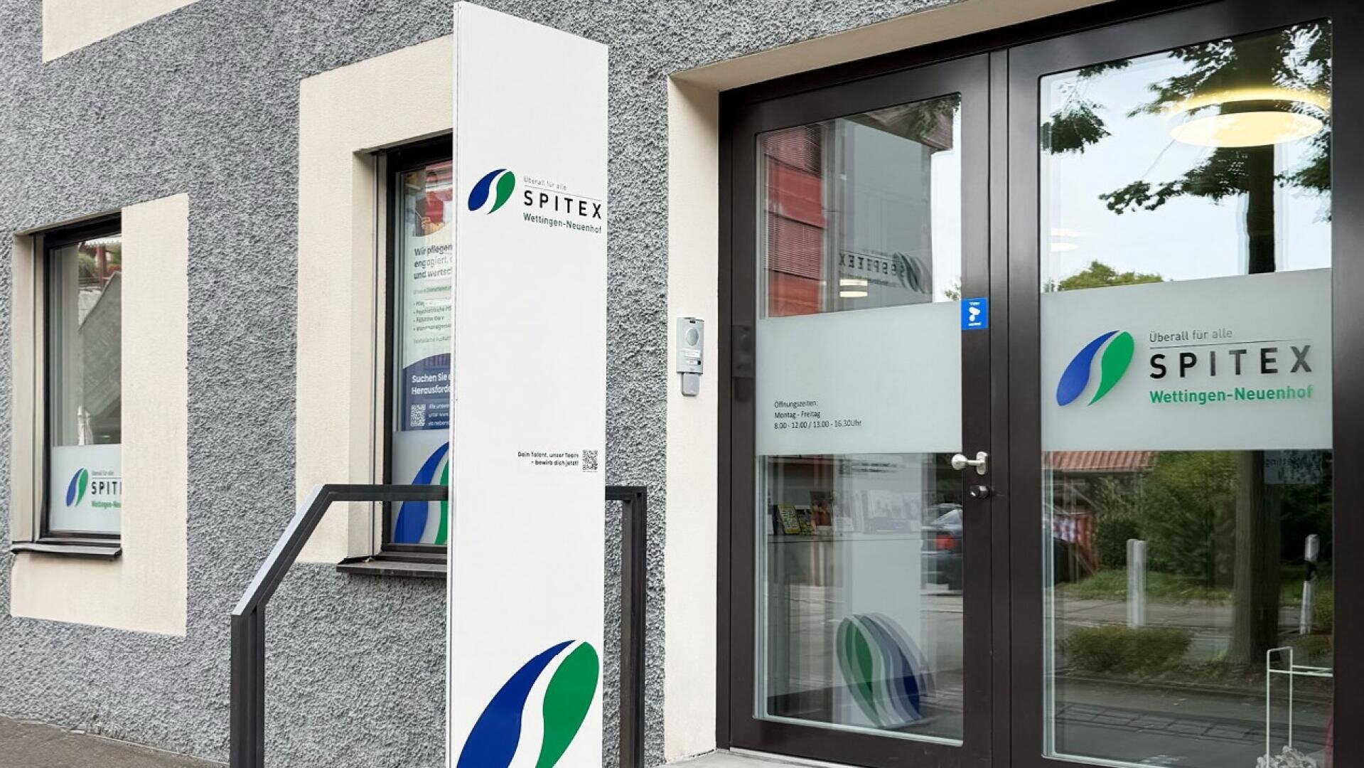 Eingang eines modernen Gebäudes mit großen Glastüren und -fenstern, an denen das Logo und der Name „SPITEX Wettingen-Neuenhof“ auf Schildern im Außenbereich und auf dem Glas zu sehen sind. Die Außenwände sind grau mit weißen Zierleisten.