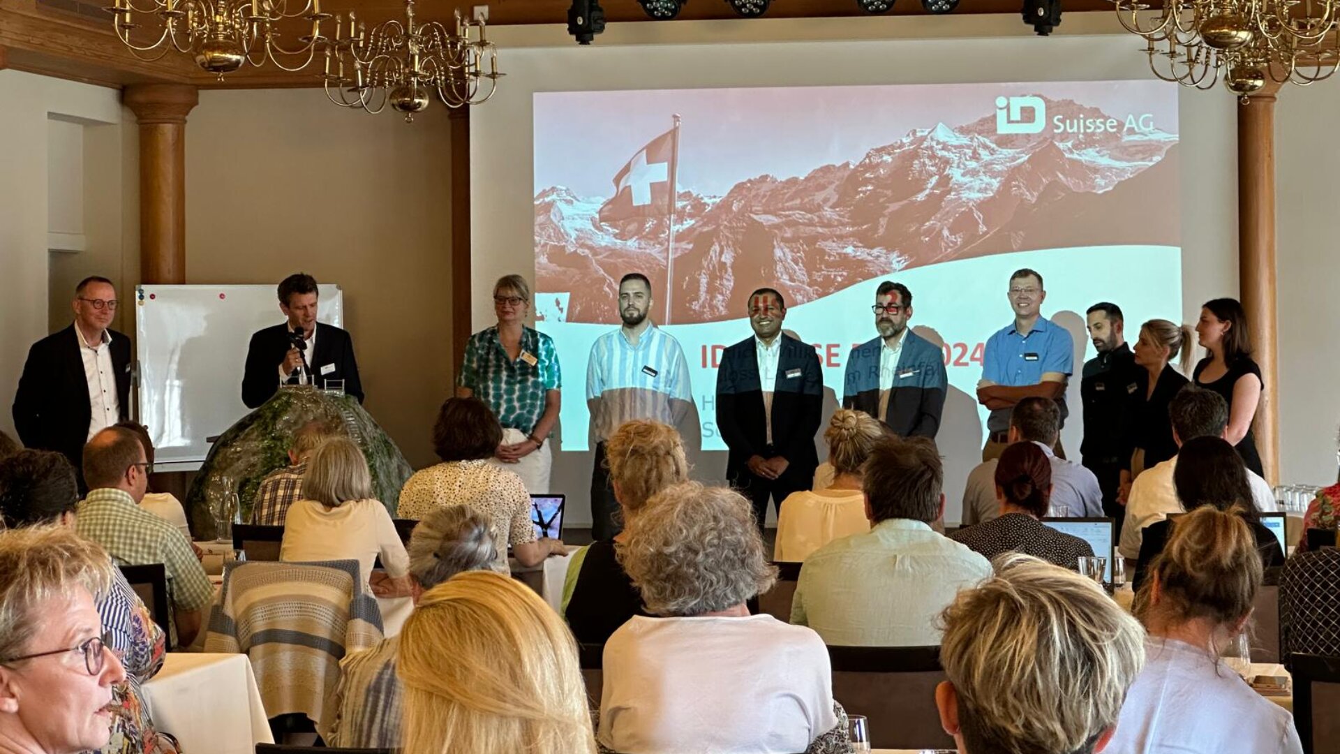 Eine Gruppe von Personen steht auf der Bühne vor einem sitzenden Publikum, hinter ihnen ein Präsentationsbildschirm, auf dem eine Schweizer Flagge, Berge und der Text ID Suisse AG zu sehen sind. Die Veranstaltung wirkt formell und professionell.