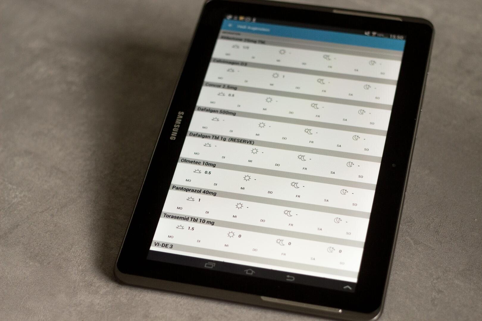 Ein Samsung-Tablet, das einen Medikamentenplan mit einer Liste von Medikamentennamen, Dosierungen und Symbolen für die Verabreichungszeiten anzeigt und auf einer grauen, strukturierten Oberfläche liegt.