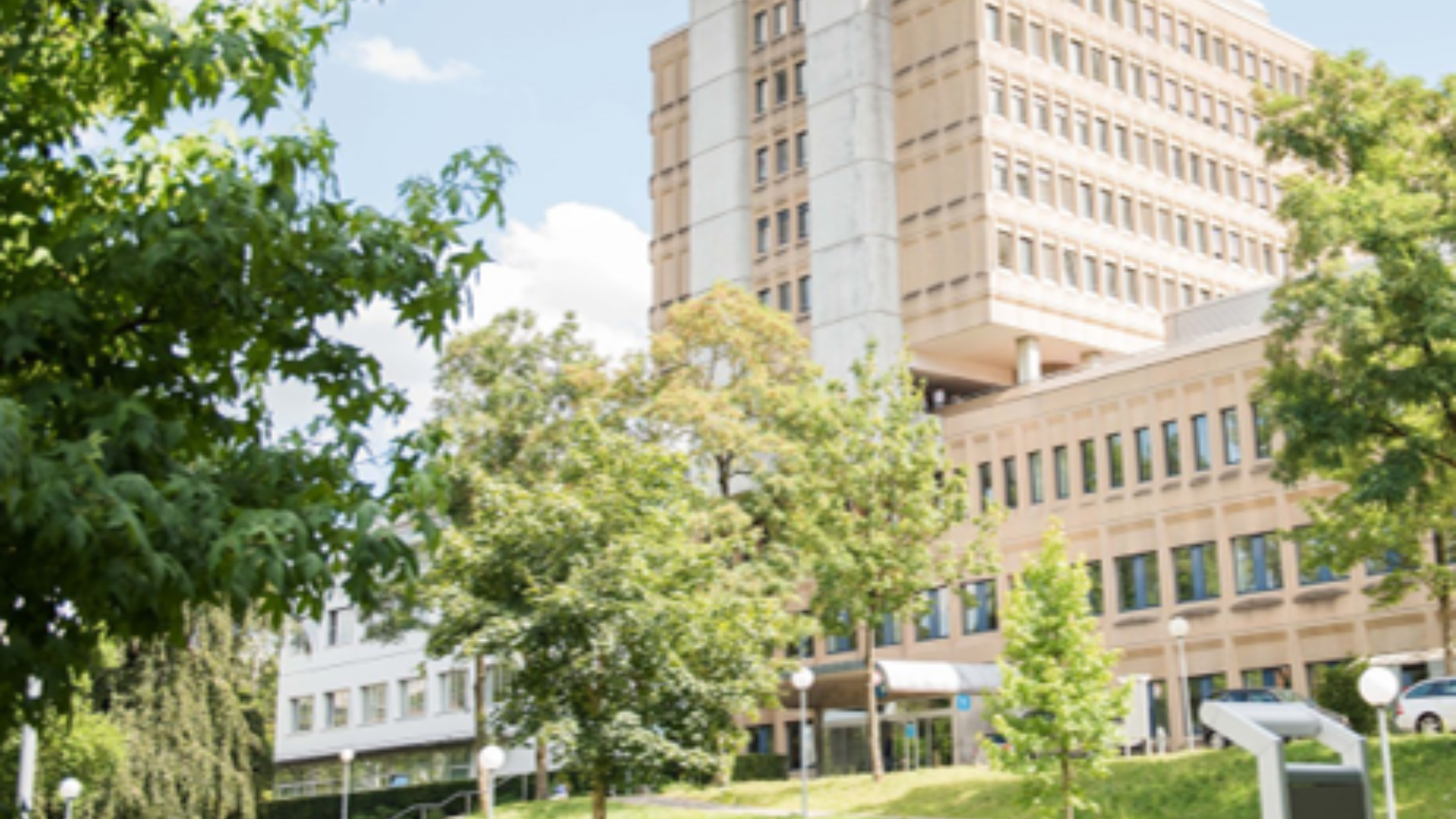Ein großes beigefarbenes Bürogebäude mit vielen Fenstern steht an einem sonnigen Tag hinter grünen Bäumen und einer Wiese. Der Himmel ist teilweise bewölkt und moderne Außenleuchten säumen den Weg.
