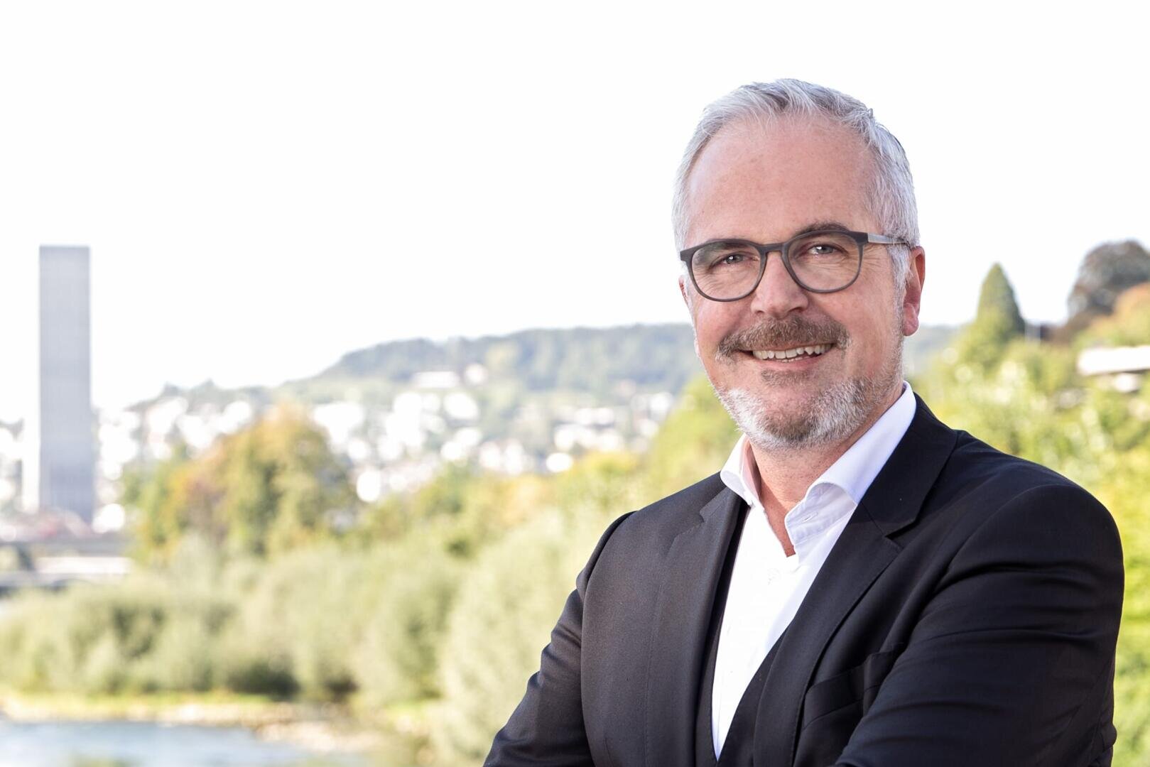 Ein lächelnder Mann mit kurzen grauen Haaren, einem Bart und einer Brille, der einen schwarzen Anzug und ein weißes Hemd trägt, steht im Freien mit unscharfen Grünflächen, Bäumen und Gebäuden im Hintergrund.