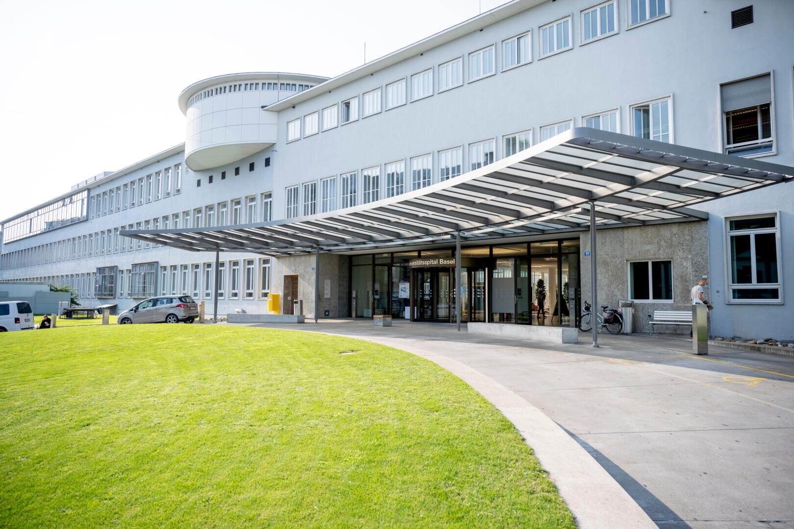 Das Bild zeigt den Eingang zu einem modernen Krankenhausgebäude mit großen Glastüren, einem Vordach aus Metall und Glas und einer geschwungenen Einfahrt, die von einem gepflegten grünen Rasen umgeben ist. In der Nähe des Eingangs befinden sich Bänke und Fahrräder.