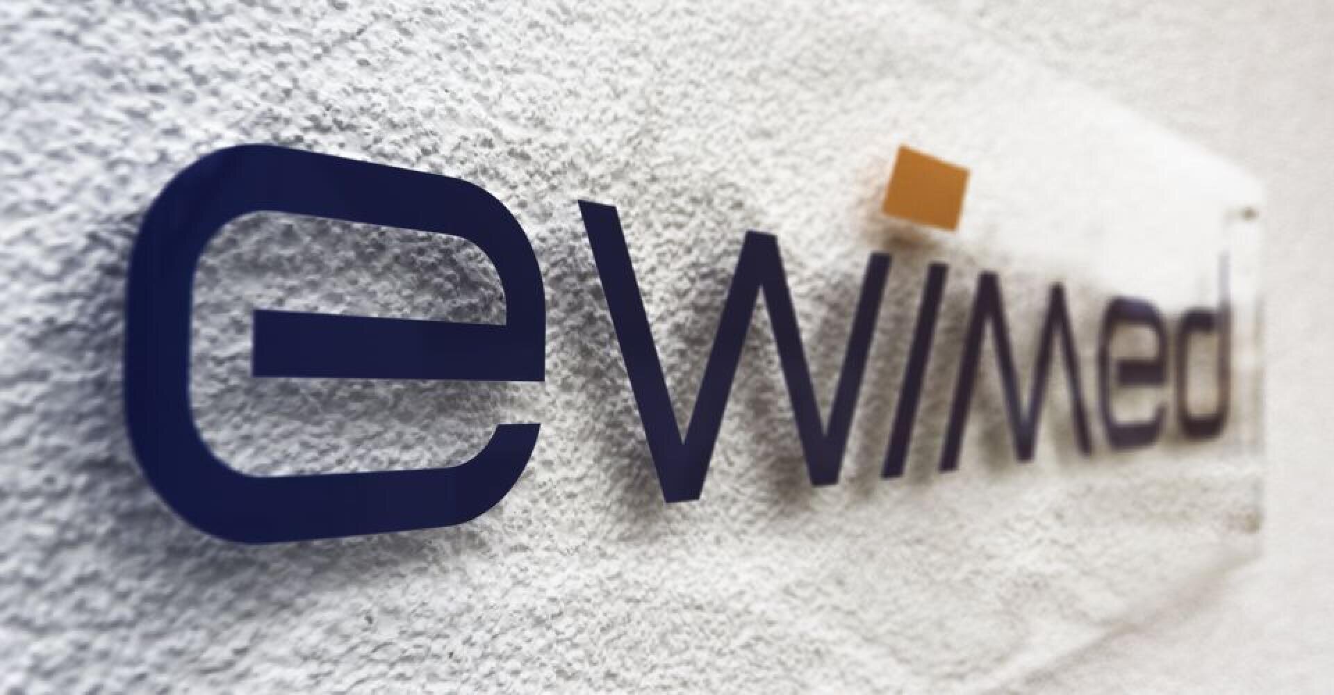 Das Firmenlogo von eWimed in dunkelblauer Schrift mit einem kleinen orangefarbenen Quadrat über dem Buchstaben i, angebracht an einer strukturierten weißen Wand.