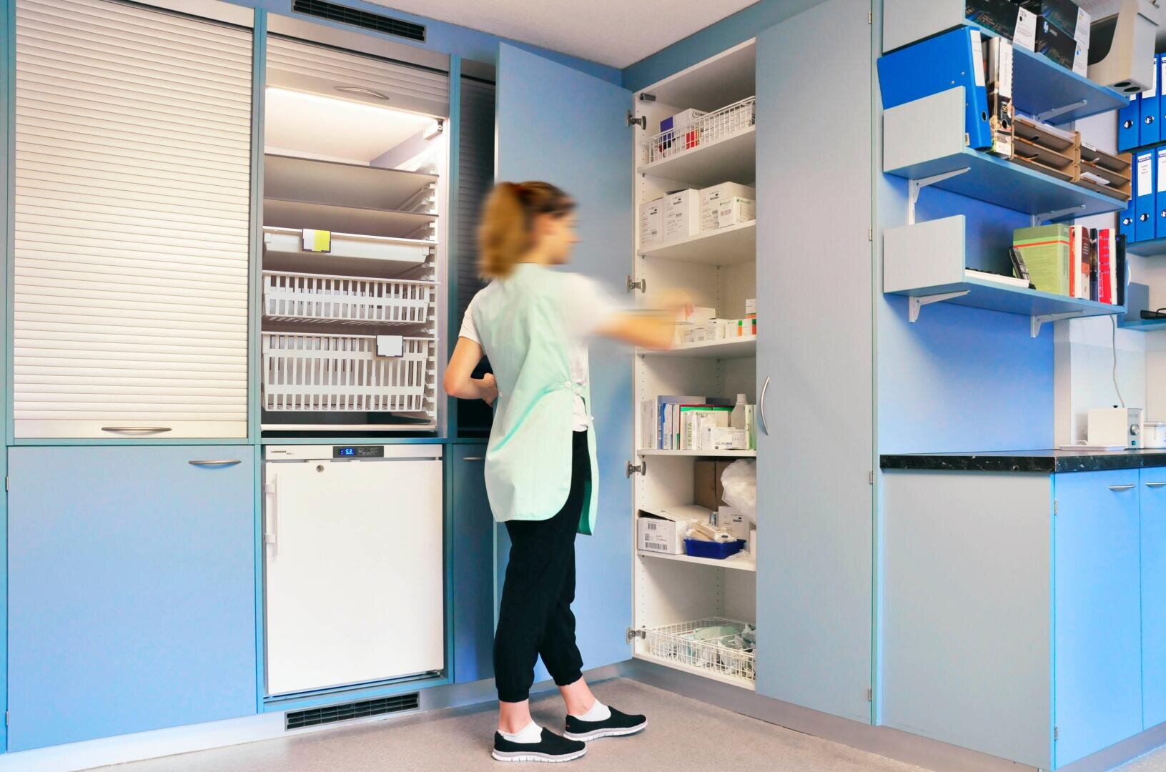 Eine Frau in medizinischer Kleidung steht in einem Krankenzimmer und greift in einen offenen Schrank, der mit medizinischem Material und Medikamenten gefüllt ist. Der Raum ist mit blauen Schränken, Tabletts und geordneten Regalen ausgestattet.