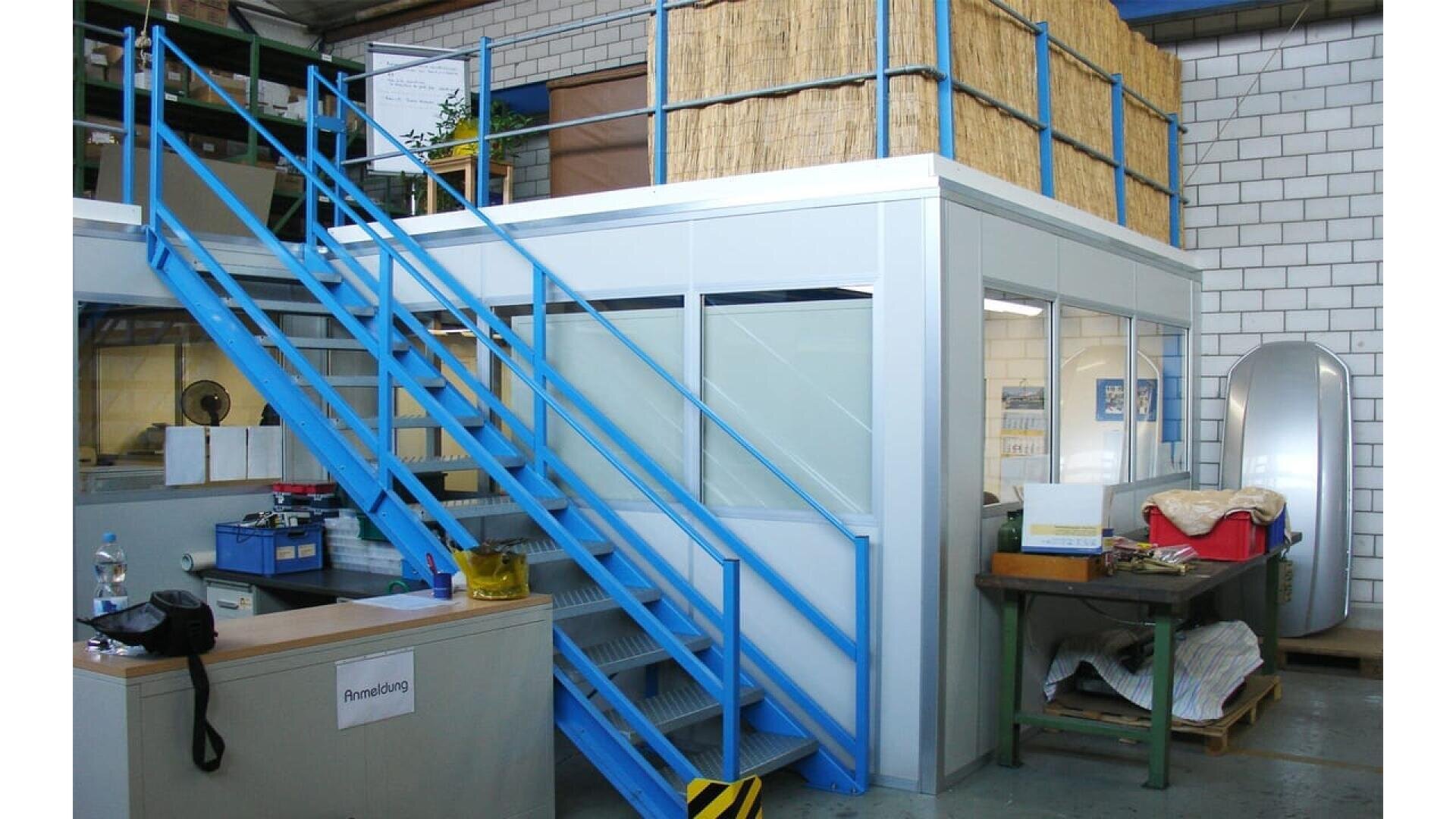 Un petit bureau aux murs de verre à l'intérieur d'un entrepôt, avec un escalier métallique bleu menant à un loft situé au-dessus. Divers objets et outils de bureau sont visibles autour de la structure.