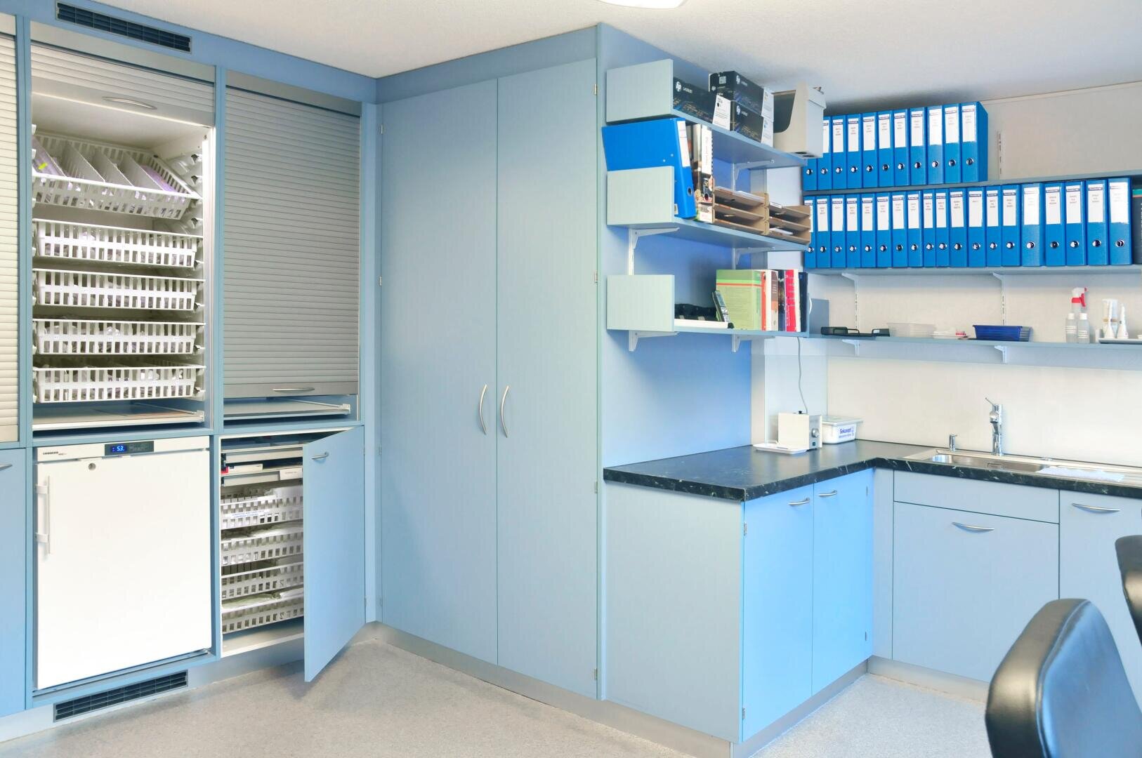 Ein sauberer, aufgeräumter Medizin- oder Laborraum mit hellblauen Schränken, Regalen mit Ordnern und Akten, einem Waschbecken und Ablagefächern, von denen einige geöffnet werden können, um Geräte und Zubehör zu zeigen.