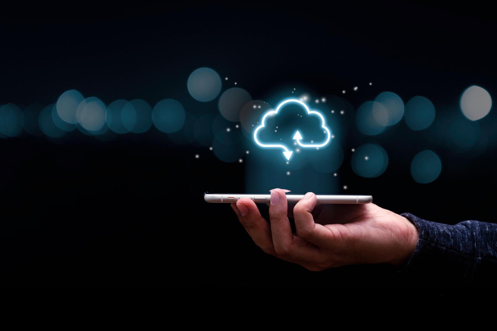 Eine Hand hält ein Smartphone mit einem leuchtenden Wolkensymbol darüber, das Cloud Computing oder Daten-Upload symbolisiert. Der Hintergrund ist dunkel mit verschwommenen Lichtern, die die Wolke und das Gerät hervorheben.