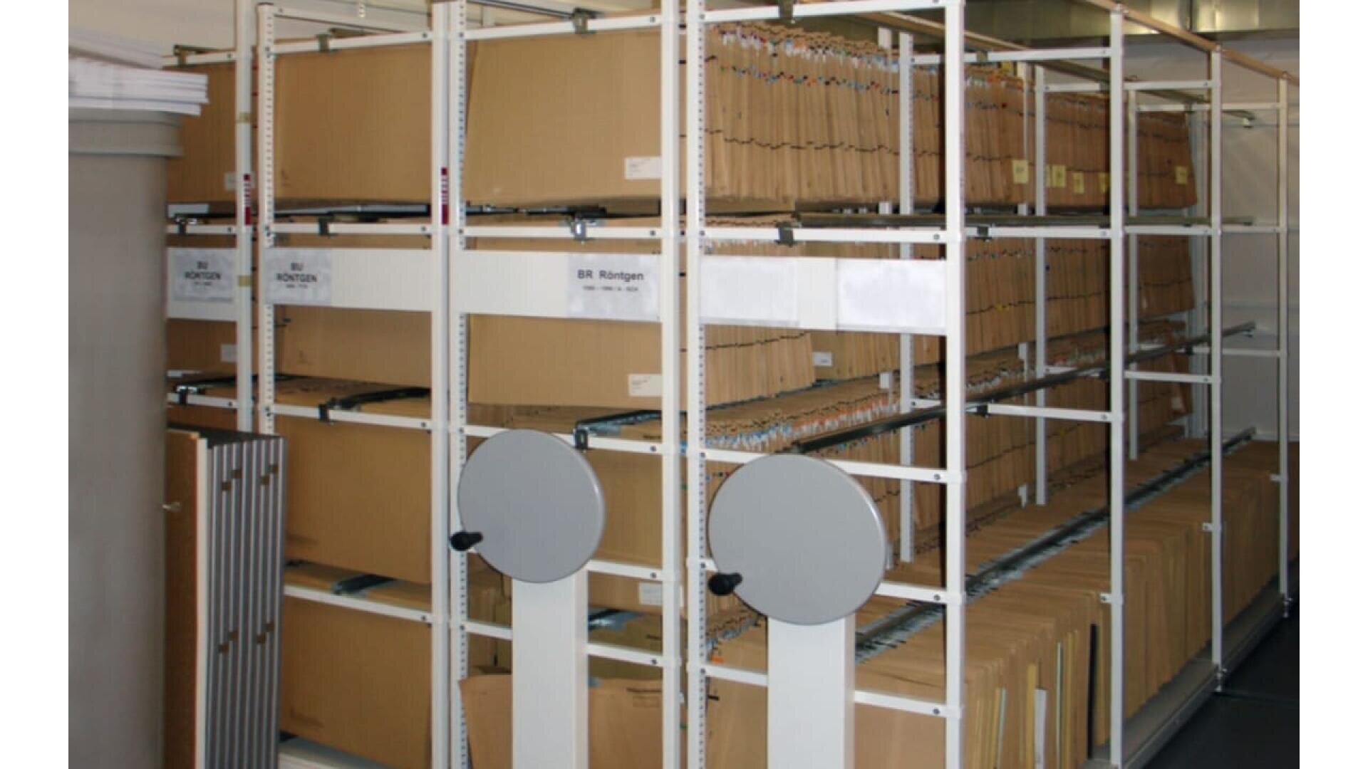 Étagères de dossiers bruns dans un rayonnage mobile, avec des poignées rondes pour déplacer les étagères. Chaque dossier est étiqueté et organisé, et contient probablement des documents ou des enregistrements. L'endroit semble être une salle d'archives ou de stockage de dossiers.